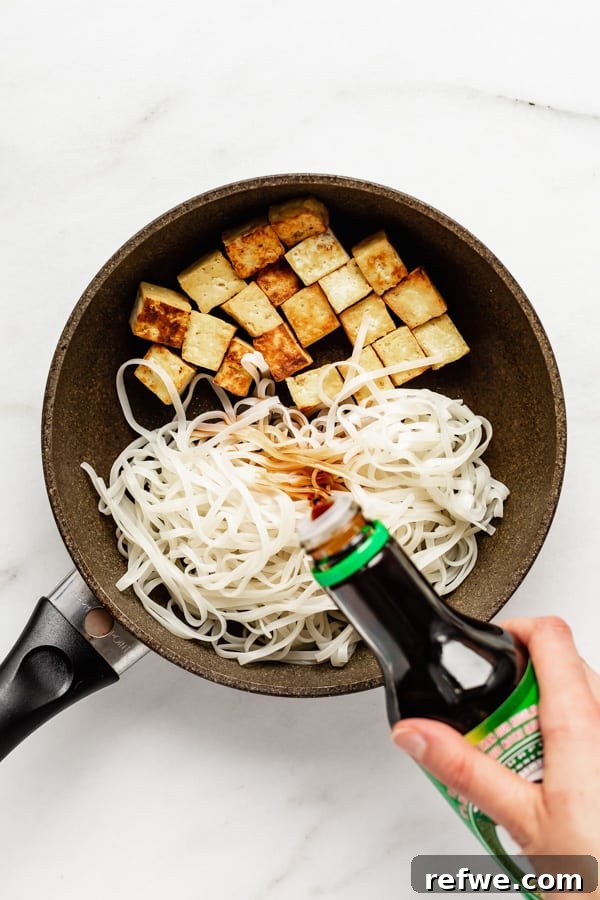 Tofu and rice noodles being stir-fried in a pan, with a hand pouring soy sauce over the mixture.