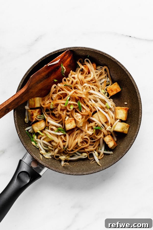 Tofu, rice noodles, and fresh bean sprouts mixed together in a pan with a wooden spoon.