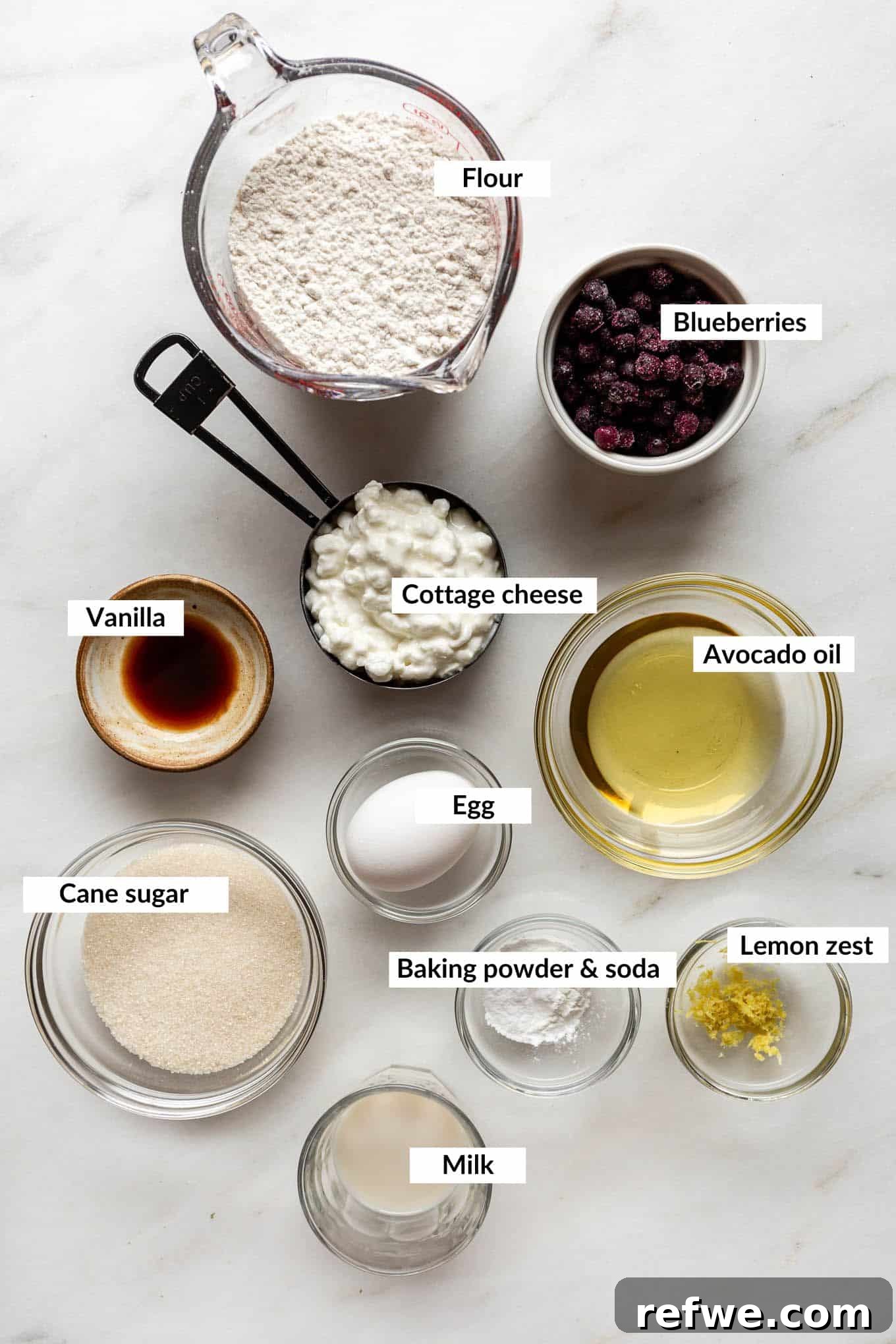 An array of fresh ingredients including cottage cheese, blueberries, flour, milk, and eggs, laid out for making blueberry cottage cheese muffins.