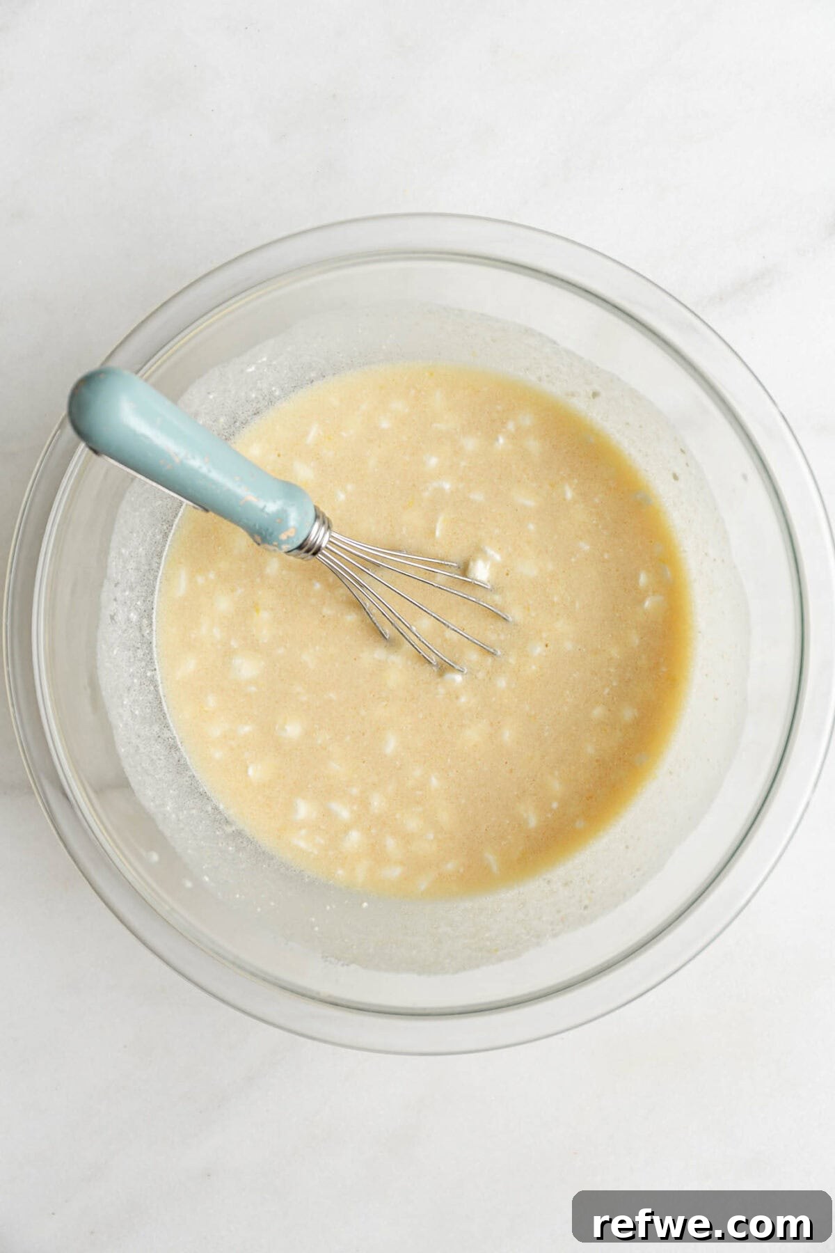 The wet ingredients for blueberry cottage cheese muffins, including sugar and lemon zest, being whisked in a clear mixing bowl with a blue whisk.