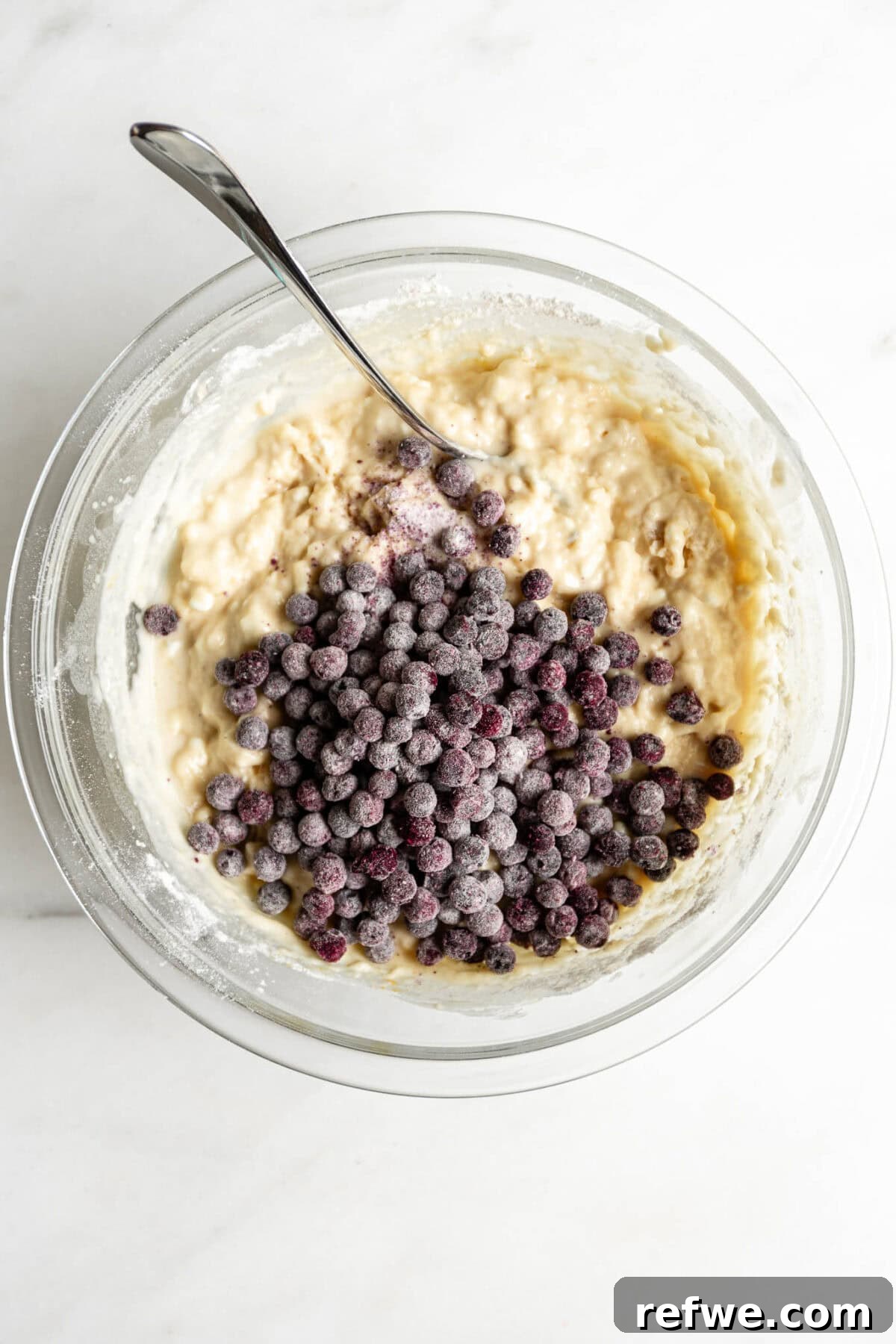 Blueberry muffin batter with a generous pile of fresh blueberries ready to be folded in, sitting in a clear mixing bowl.