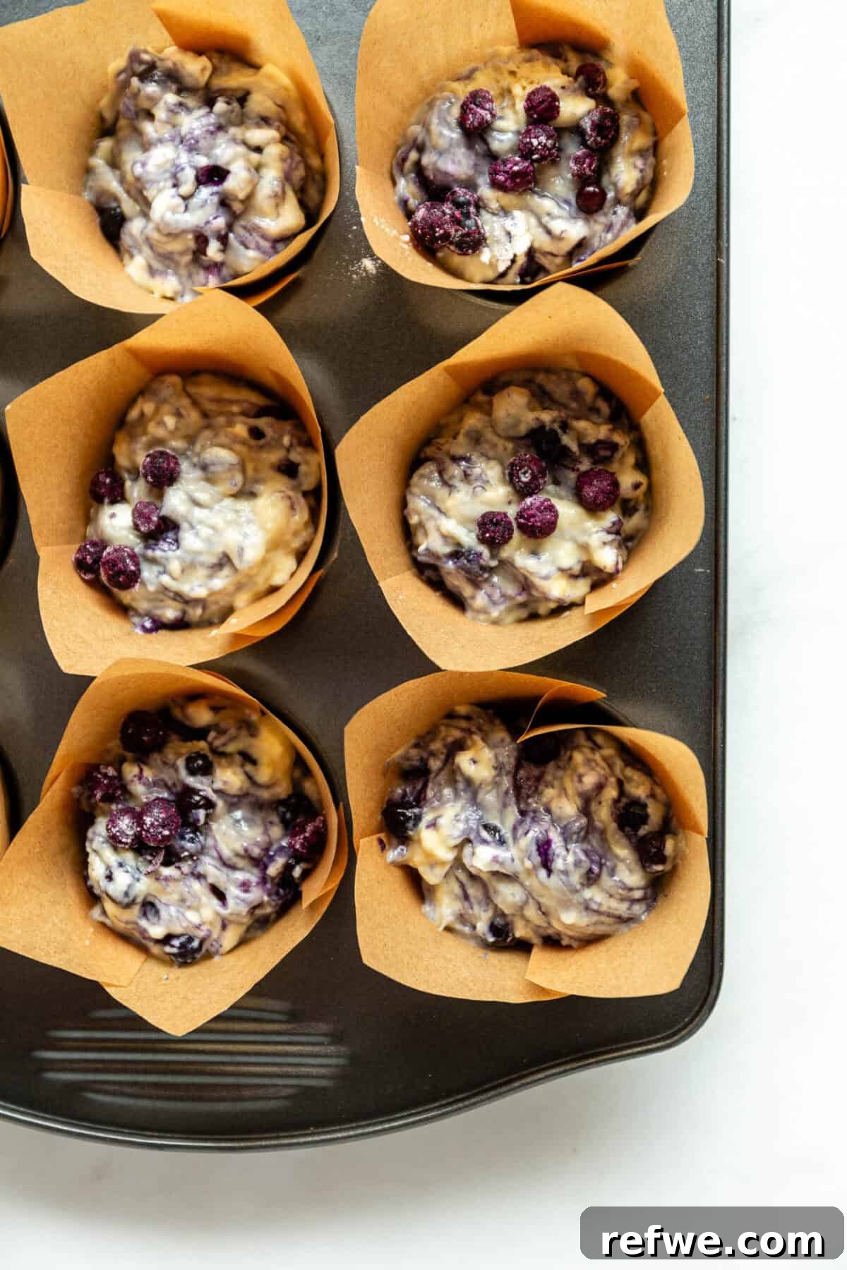 Blueberry muffin batter spooned into a muffin pan lined with paper cups, ready to be baked.