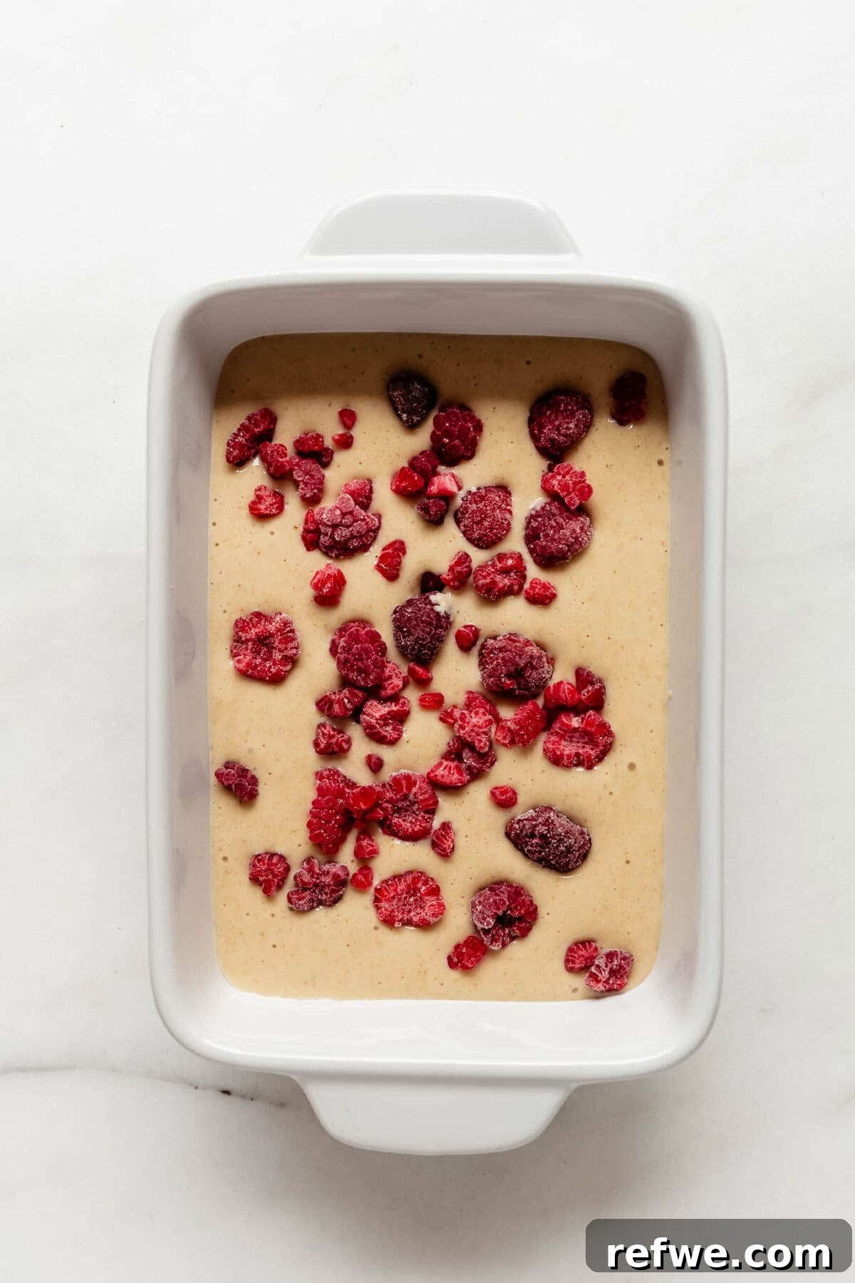 Peak Performance Pancake Bowl 7 The pancake bowl batter, now in a white baking dish, is being topped with frozen raspberries, preparing it for the baking process.