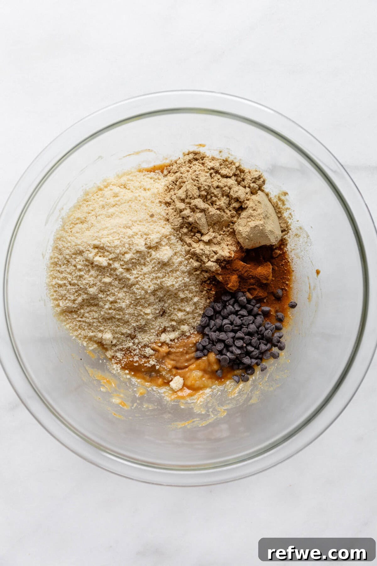 Dry ingredients, including almond flour, protein powder, spices, and chocolate chips, added to the wet mixture in a bowl.