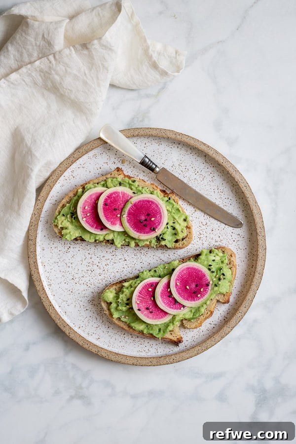 Sunrise Avocado Toast with Ruby Radish Slices 3 Two slices of smashed avocado toast, richly spread with avocado and adorned with thinly sliced watermelon radish and a sprinkle of sesame seeds, ready to eat.