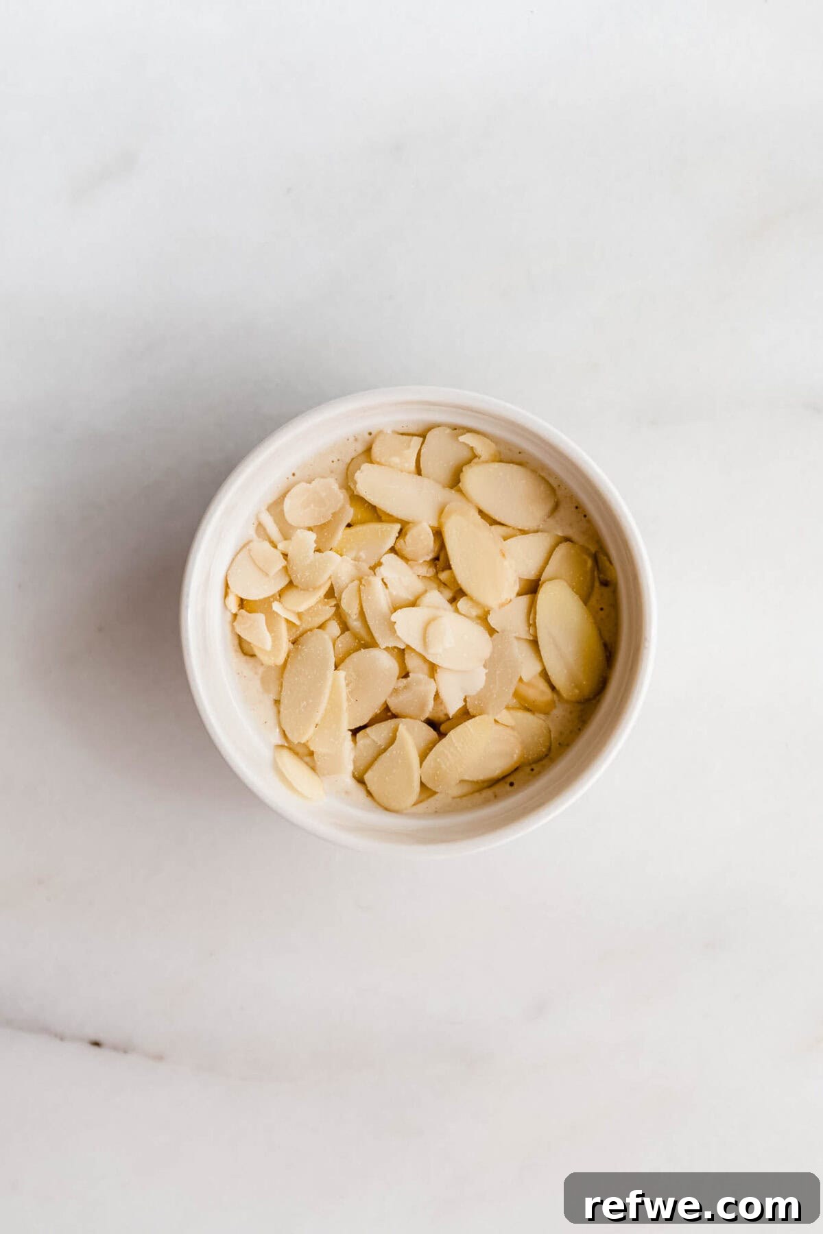 Assembled almond croissant baked oats in ramekins topped with sliced almonds, ready for baking.