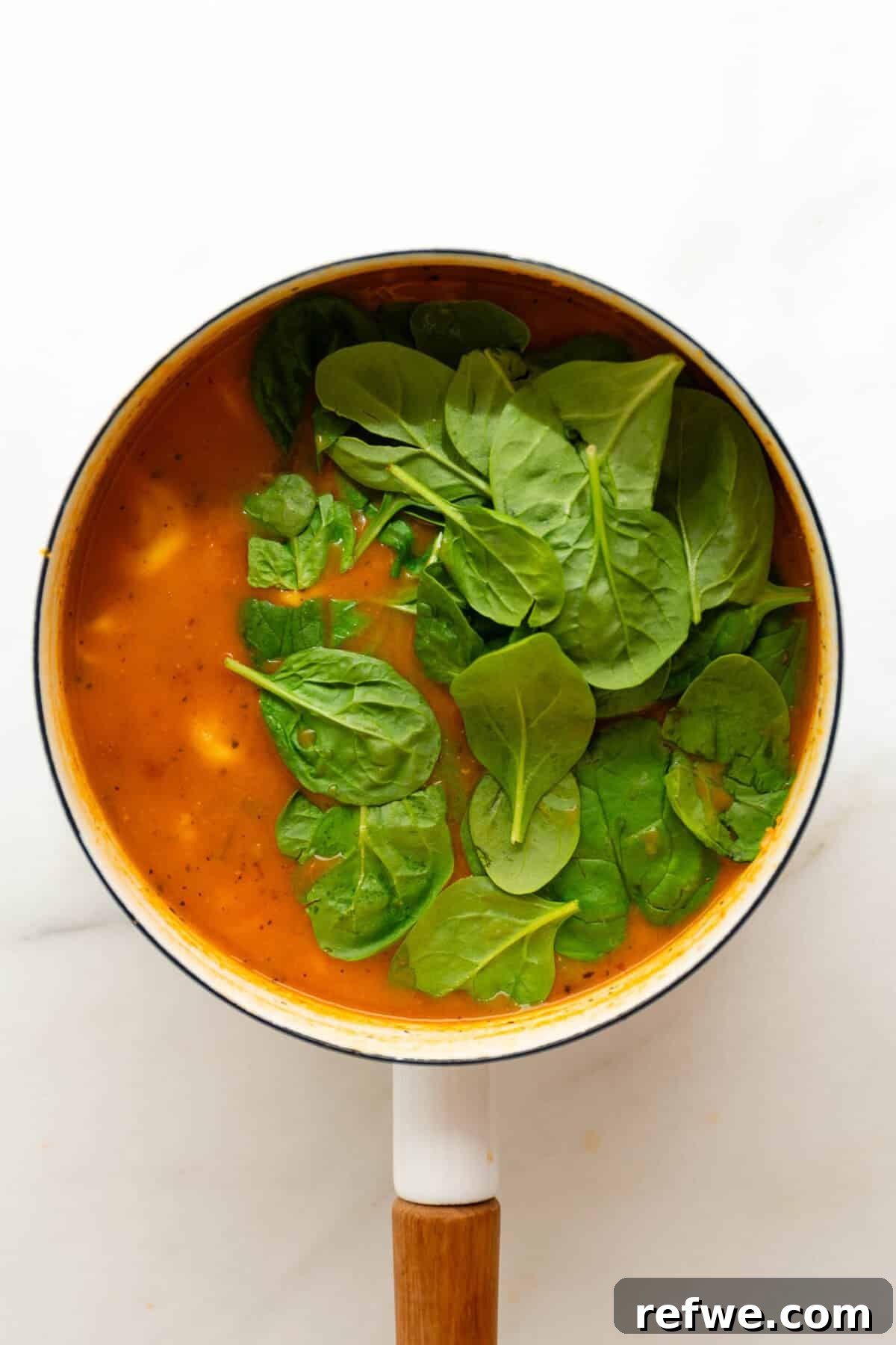Velvety Tuscan Tortellini Soup 10 Fresh spinach leaves being stirred into a pot of creamy Tuscan tortellini soup, beginning to wilt.