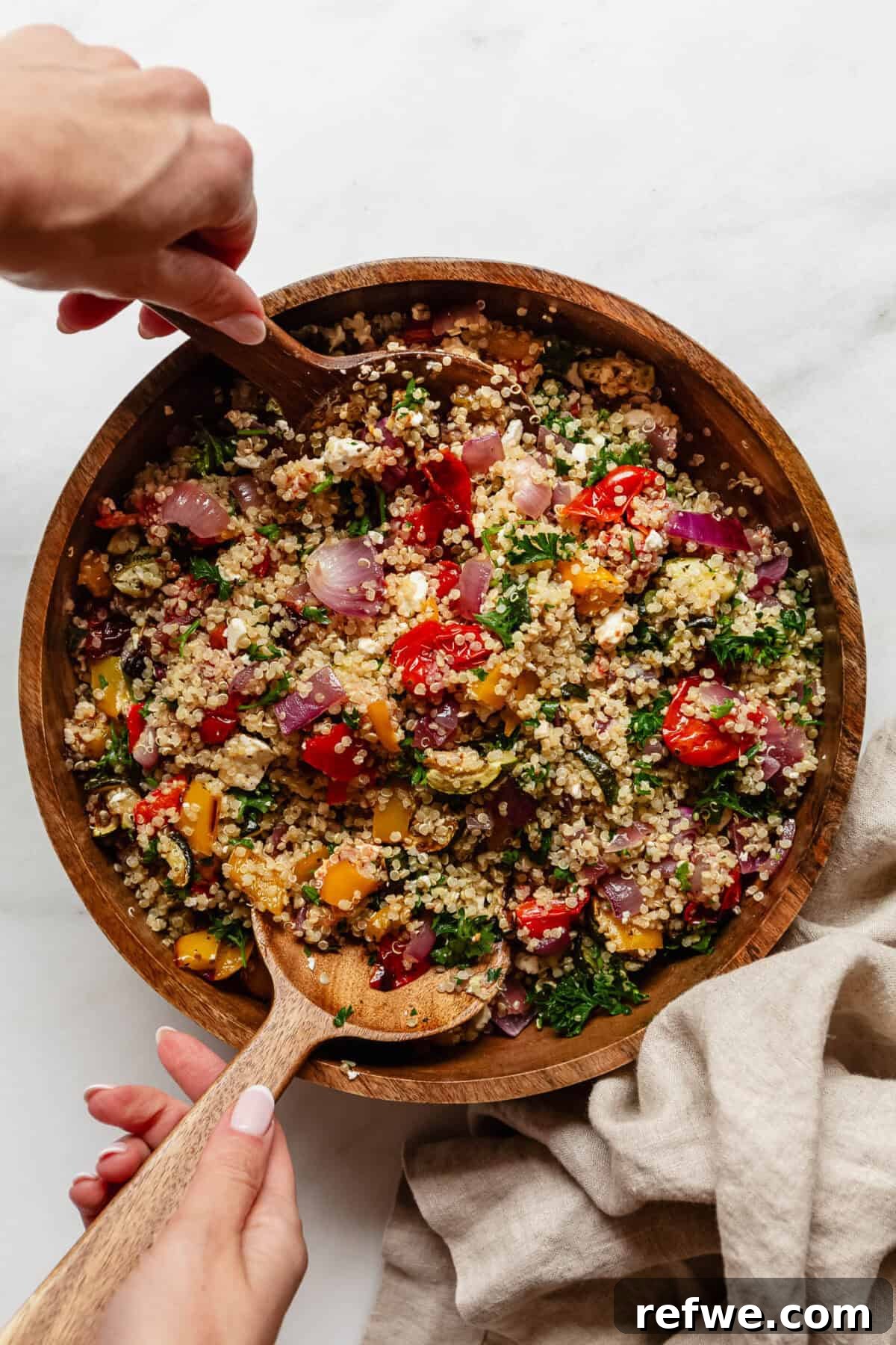 Vibrant Roasted Vegetable Quinoa Salad 7 Two hands using wooden serving spoons to gently toss the roasted vegetable quinoa salad in a large wooden bowl, ensuring all ingredients are well mixed.