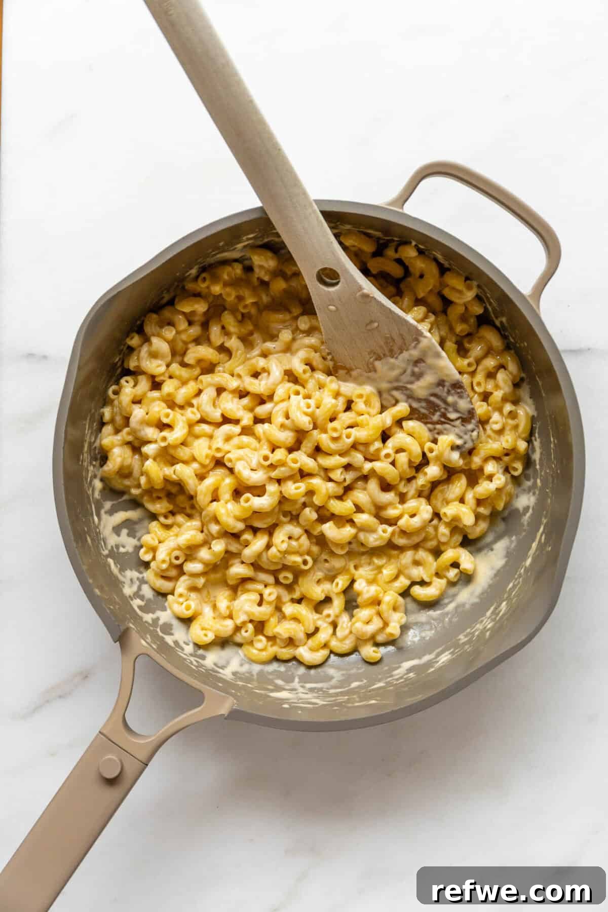 15-Minute Cottage Cheese Mac & Cheese Delight 7 A beige pan filled with rich, creamy cottage cheese mac and cheese, being stirred with a wooden spoon, indicating it's ready to serve.