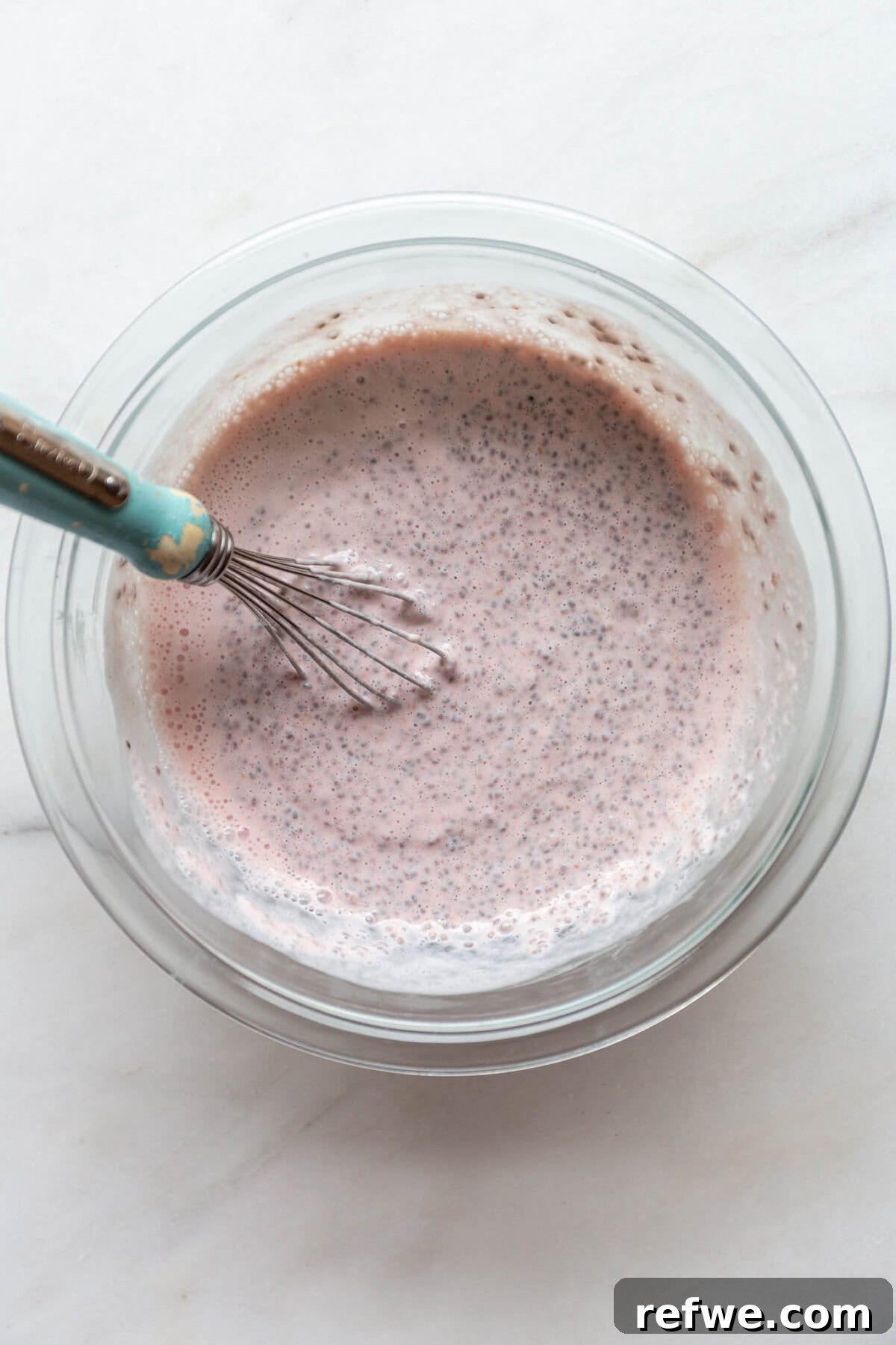 Supercharged Protein Chia Pudding 7 Pink chia pudding in a clear mixing bowl with a blue whisk.
