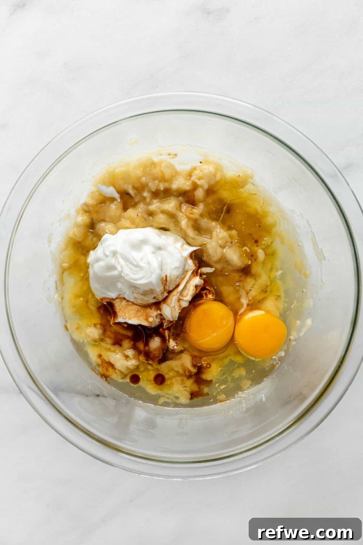 Mashed banana, cracked eggs, and creamy Greek yogurt being combined in a clear mixing bowl, ready for the next step.