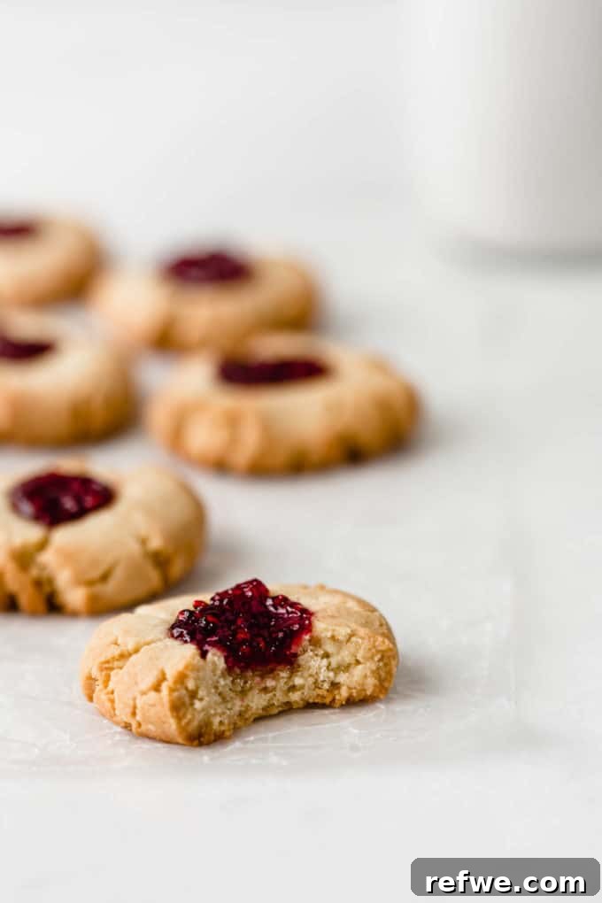 A delectable vegan raspberry thumbprint cookie with a small bite taken out, resting elegantly on a pristine marble counter, showcasing its golden crust and vibrant red jam filling.