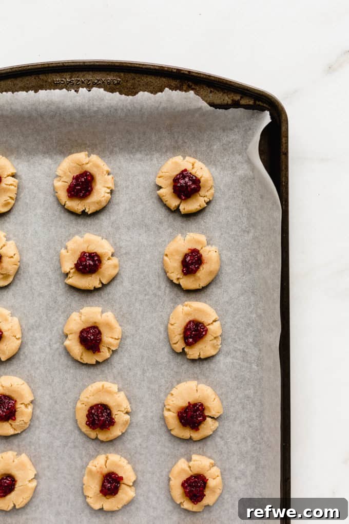 Unbaked thumbprint cookies on a baking sheet, now filled with vibrant raspberry jam, prepared for the oven.