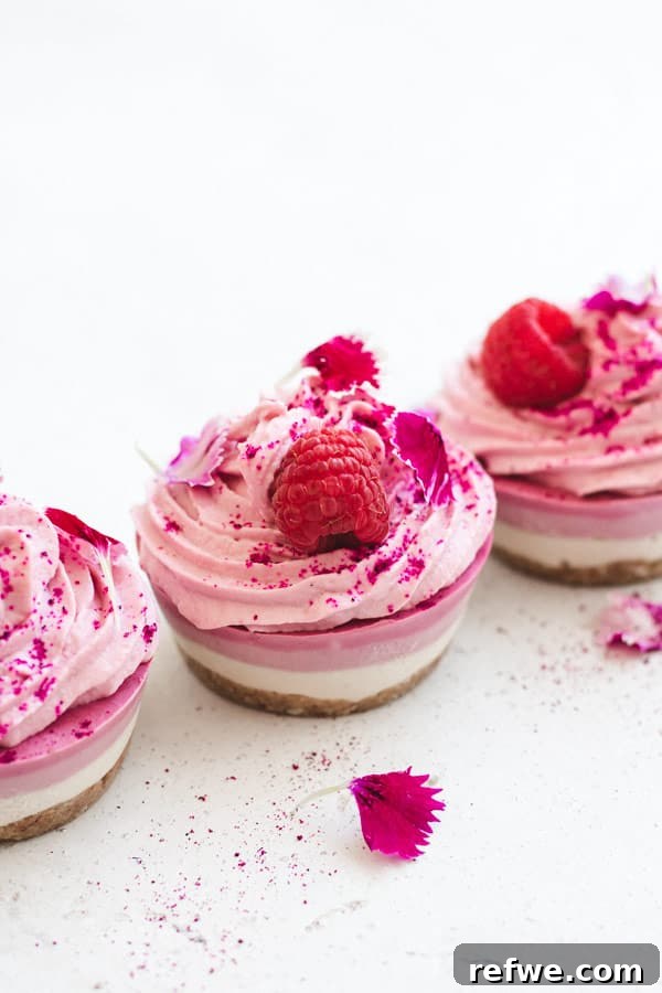 Three individual raw vegan raspberry cheesecakes arranged on a white stone surface, topped with fresh berries and a hint of green leaf.
