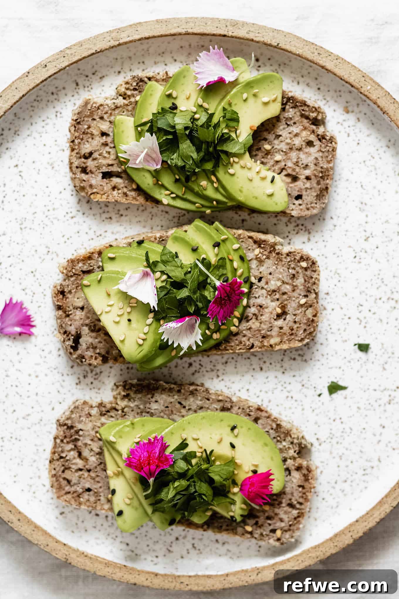 Wholesome Chia Buckwheat Loaf 2 Three slices of perfectly toasted avocado toast on a white plate, adorned with delicate pink flower petals, showcasing the versatility of Buckwheat Chia Bread.
