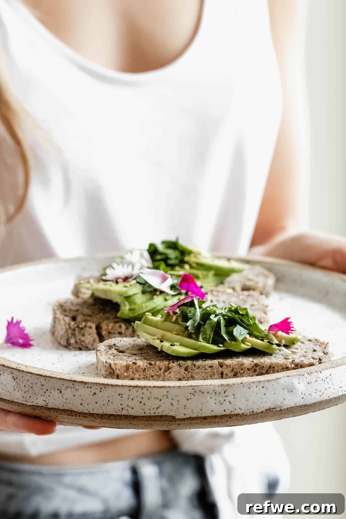 Wholesome Chia Buckwheat Loaf 10 A person gently holds a white plate featuring a slice of avocado toast, beautifully adorned with pink flower petals, highlighting the appealing presentation of the buckwheat chia bread.