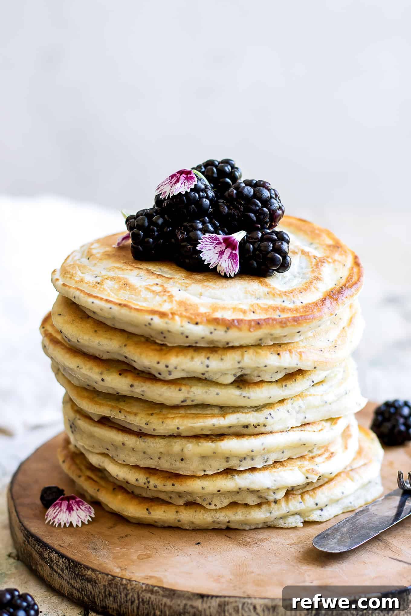 A generous stack of golden-brown lemon poppy seed spelt pancakes adorned with fresh, juicy blackberries and a luscious drizzle of golden maple syrup, ready to be enjoyed.