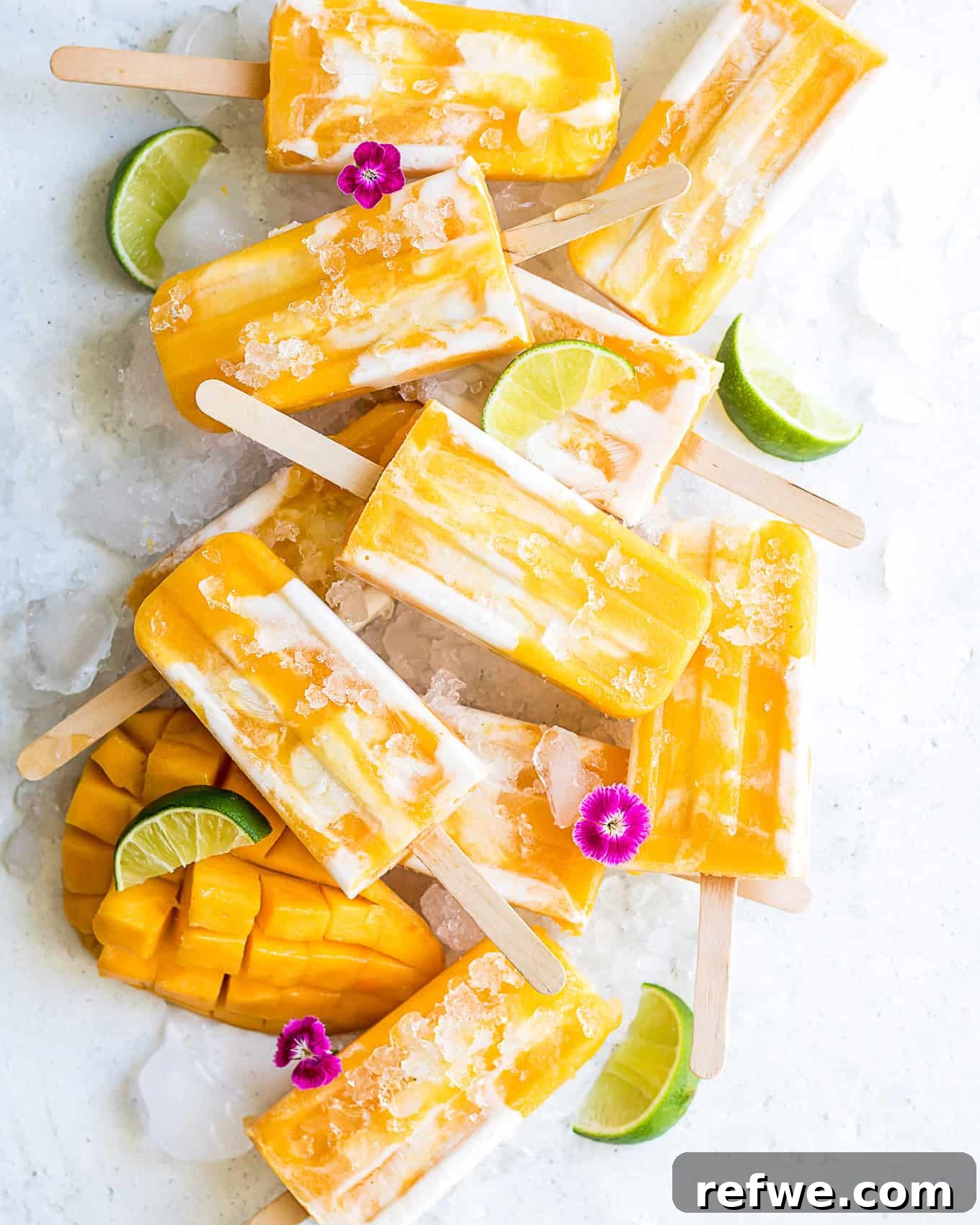 Creamy Mango Lassi Pops 2 Vibrant vegan mango lassi popsicles artfully arranged on a bed of ice, garnished with fresh mango slices, bright green limes, and delicate pink flowers, highlighting their refreshing appeal.