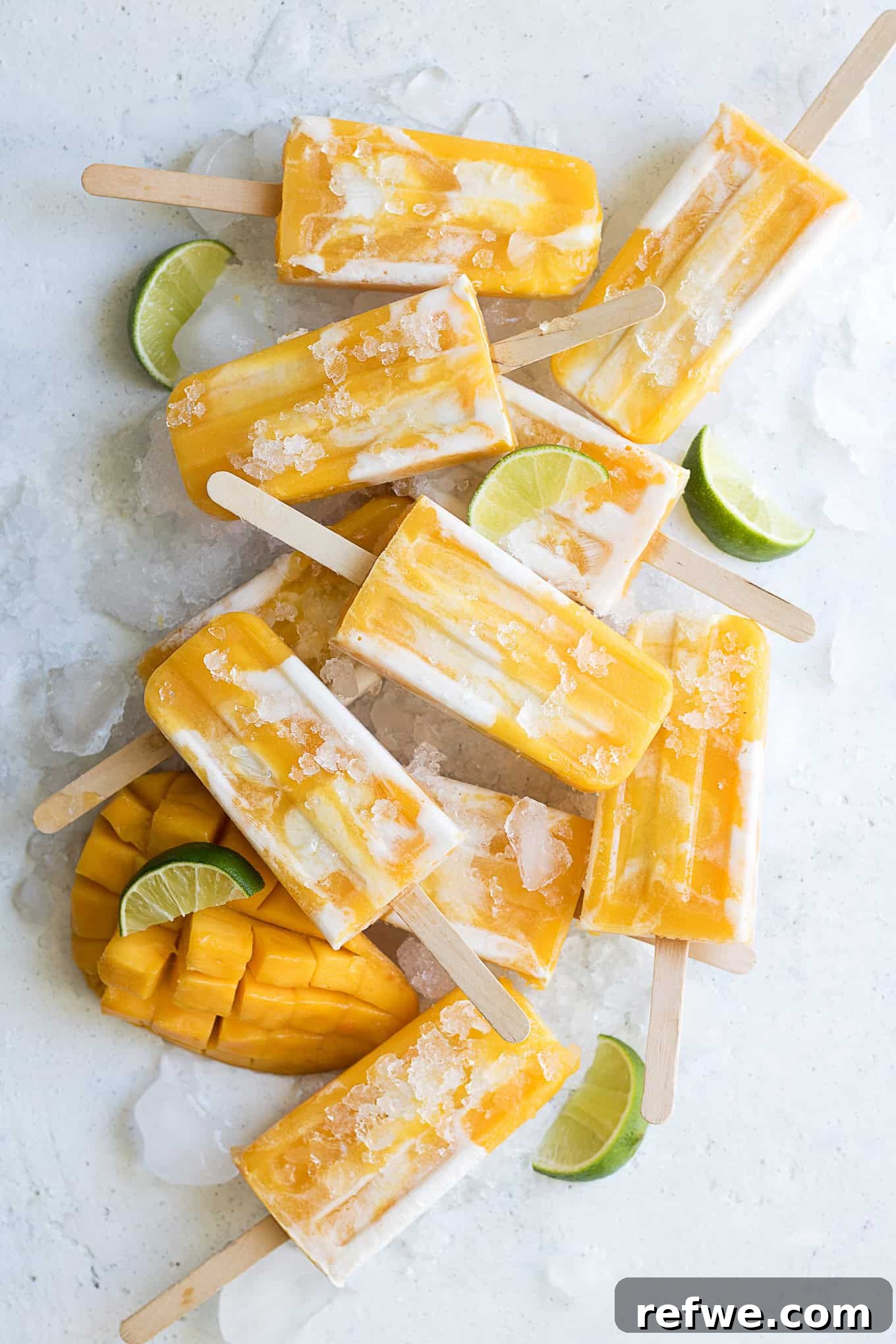 Creamy Mango Lassi Pops 5 A close-up of a few mango lassi popsicles, standing upright on ice, with fresh mango slices and limes in the background, emphasizing their refreshing qualities.