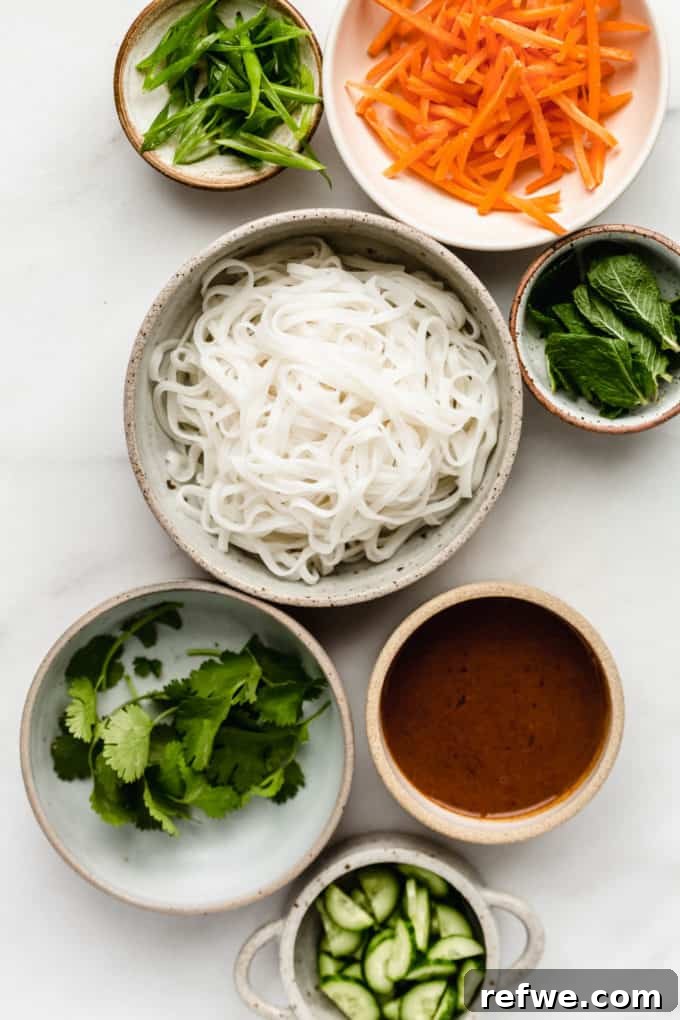Spring Roll Power Bowls with Zesty Almond Butter Sauce 3 Bowls filled with uncooked rice noodles, shredded carrots, sliced cucumbers, and fresh herbs, ready for assembly.