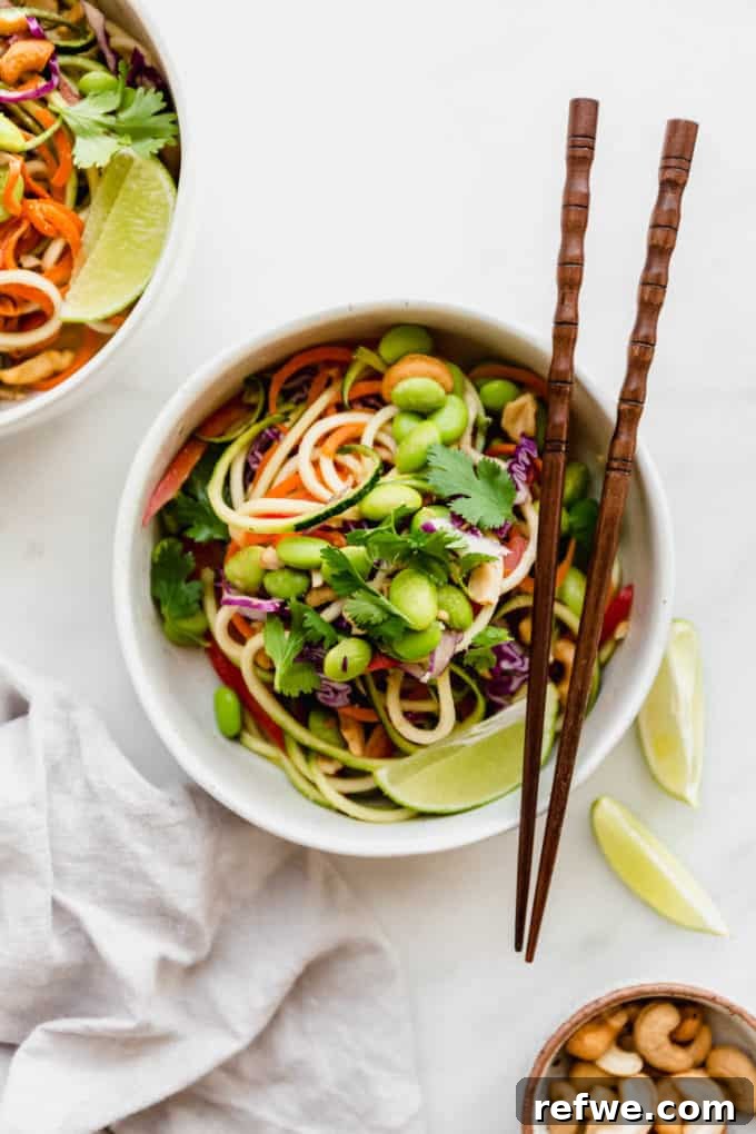 Vibrant Raw Pad Thai Noodles 2 A beautifully arranged bowl of Raw Pad Thai, featuring vibrant zucchini noodles, shredded carrots, purple cabbage, and edamame, garnished with fresh cilantro and crushed cashews, presented with elegant wooden chopsticks on a pristine white surface.