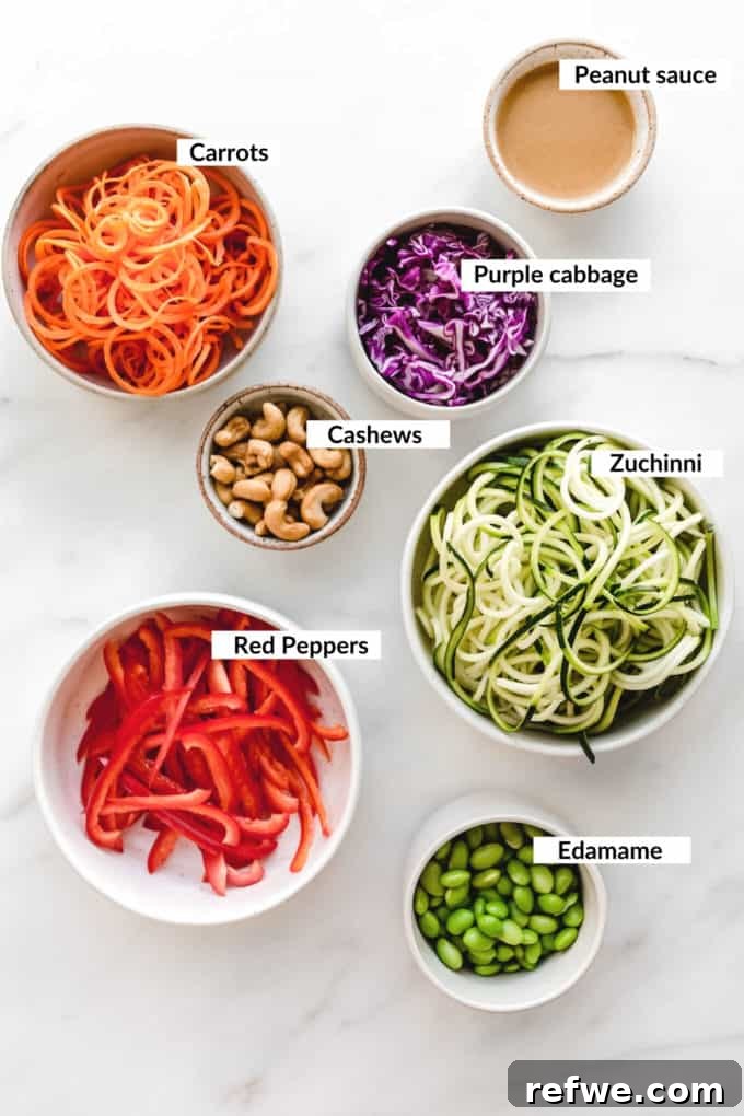 Vibrant Raw Pad Thai Noodles 3 A beautifully composed flat lay of fresh Raw Pad Thai ingredients, including bright green zucchini, orange carrots, red bell peppers, purple cabbage, shelled edamame, and whole cashews, with a small bowl of rich peanut sauce ready for mixing, all artfully arranged on a light background.