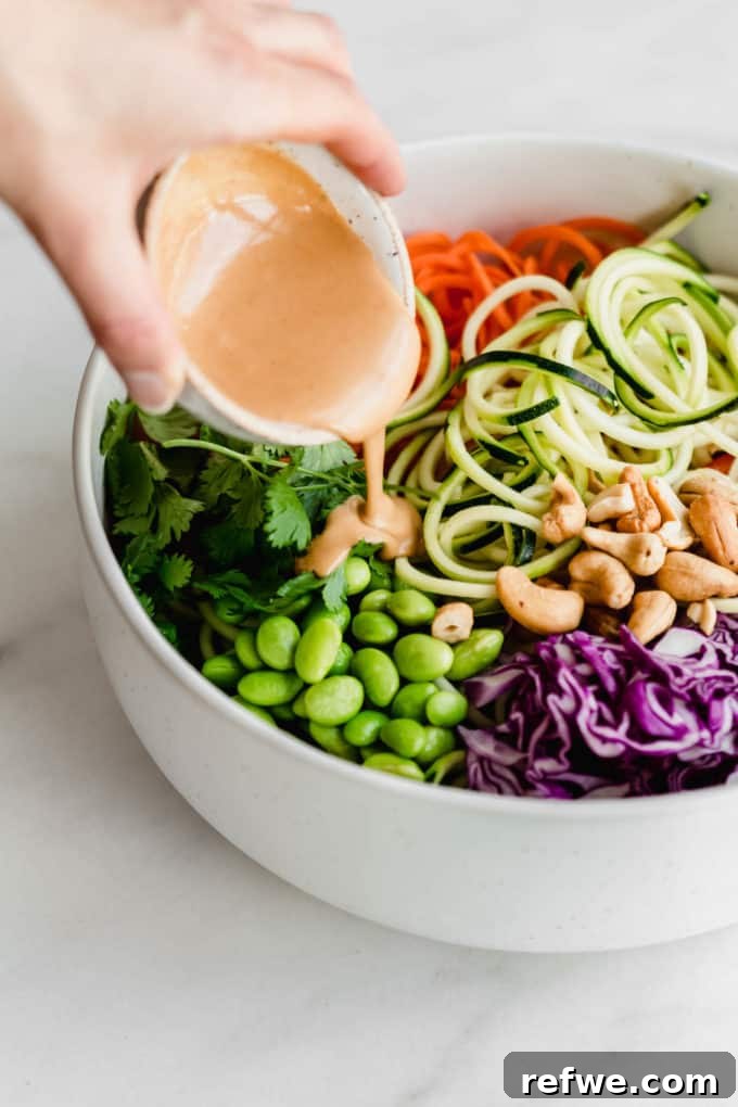 Vibrant Raw Pad Thai Noodles 5 A hand gently pouring a rich, creamy peanut sauce over the vibrant mixed vegetables in a white bowl, ensuring even coating for the Raw Pad Thai salad.