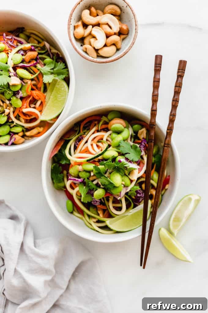 Vibrant Raw Pad Thai Noodles 6 Two pristine white bowls filled with a vibrant Raw Pad Thai, showcasing neatly spiralized zucchini noodles generously topped with bright green edamame and fresh green cilantro, beautifully presented on a light wooden surface.