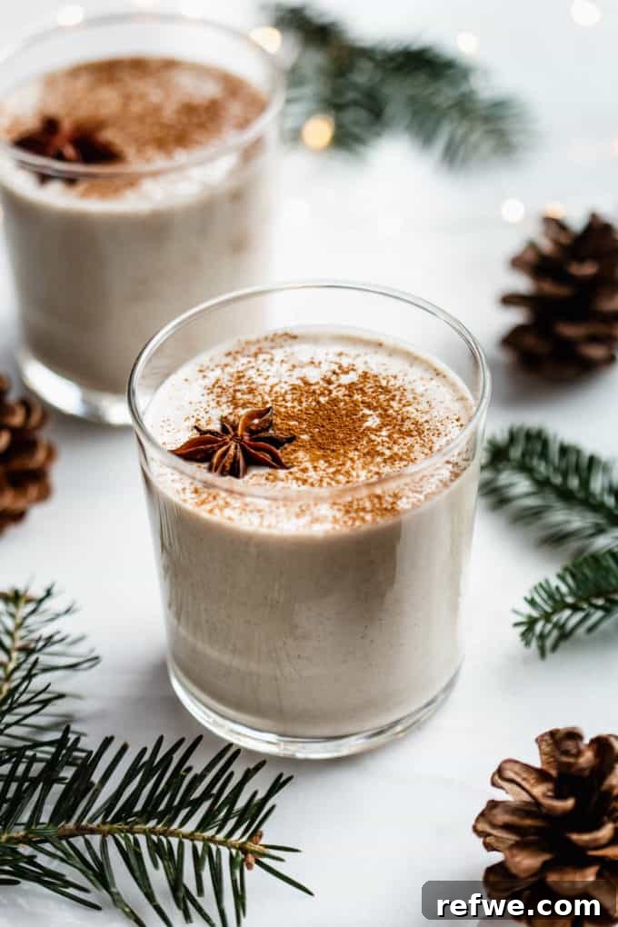 A glass of vegan eggnog on a white counter surrounded by pinecones and pine leaves, evoking a festive holiday mood.