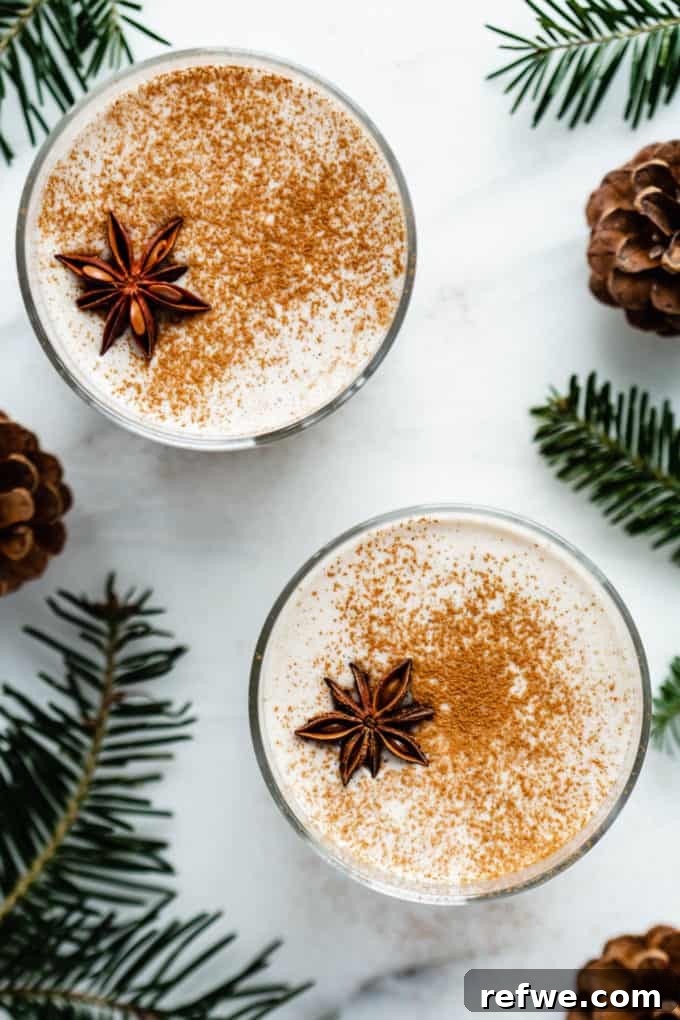 A top-down view of two festive glasses of vegan eggnog, garnished and surrounded by pine leaves on a white counter.