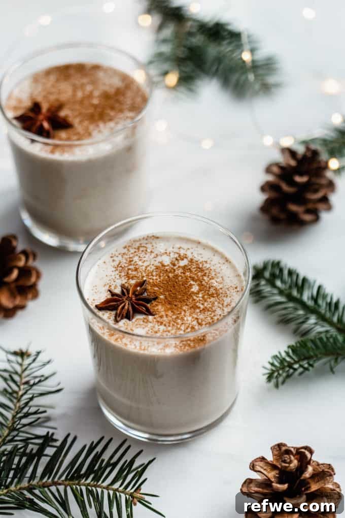 Two cups of homemade vegan eggnog topped with grated nutmeg, with warm Christmas lights gently blurred in the background.