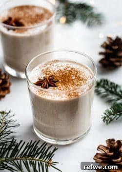 A glass of vegan eggnog on a white counter surrounded by pinecones and pine leaves