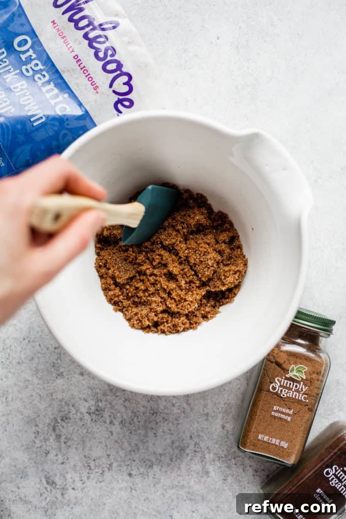 A hand mixing brown sugar and spices in a white mixing bowl, illustrating the careful blending of dry ingredients for the gingerbread cake.
