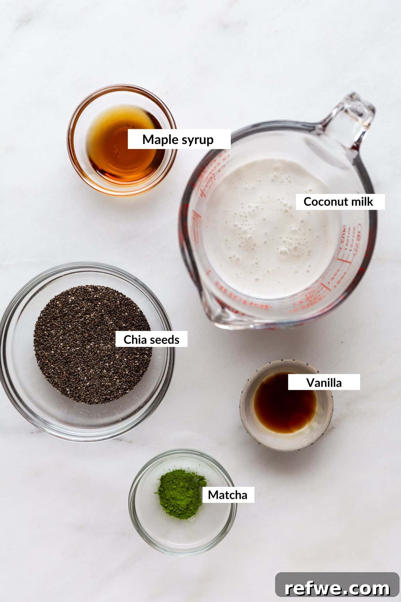 Vibrant Matcha Chia Pudding 3 A flat lay photograph showcasing the key ingredients for matcha chia pudding: a bowl of chia seeds, a container of vibrant green matcha powder, a can of full-fat coconut milk, and a bottle of maple syrup, neatly arranged on a rustic wooden surface.