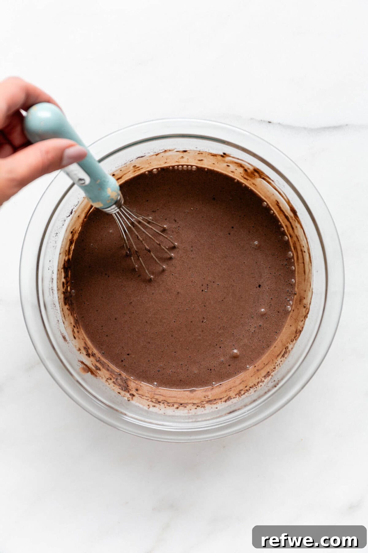 Silky Chocolate Chia Pudding 7 An un-set chocolate chia mousse mixture is being whisked in a mixing bowl using a blue whisk, showing a slightly thickened consistency.