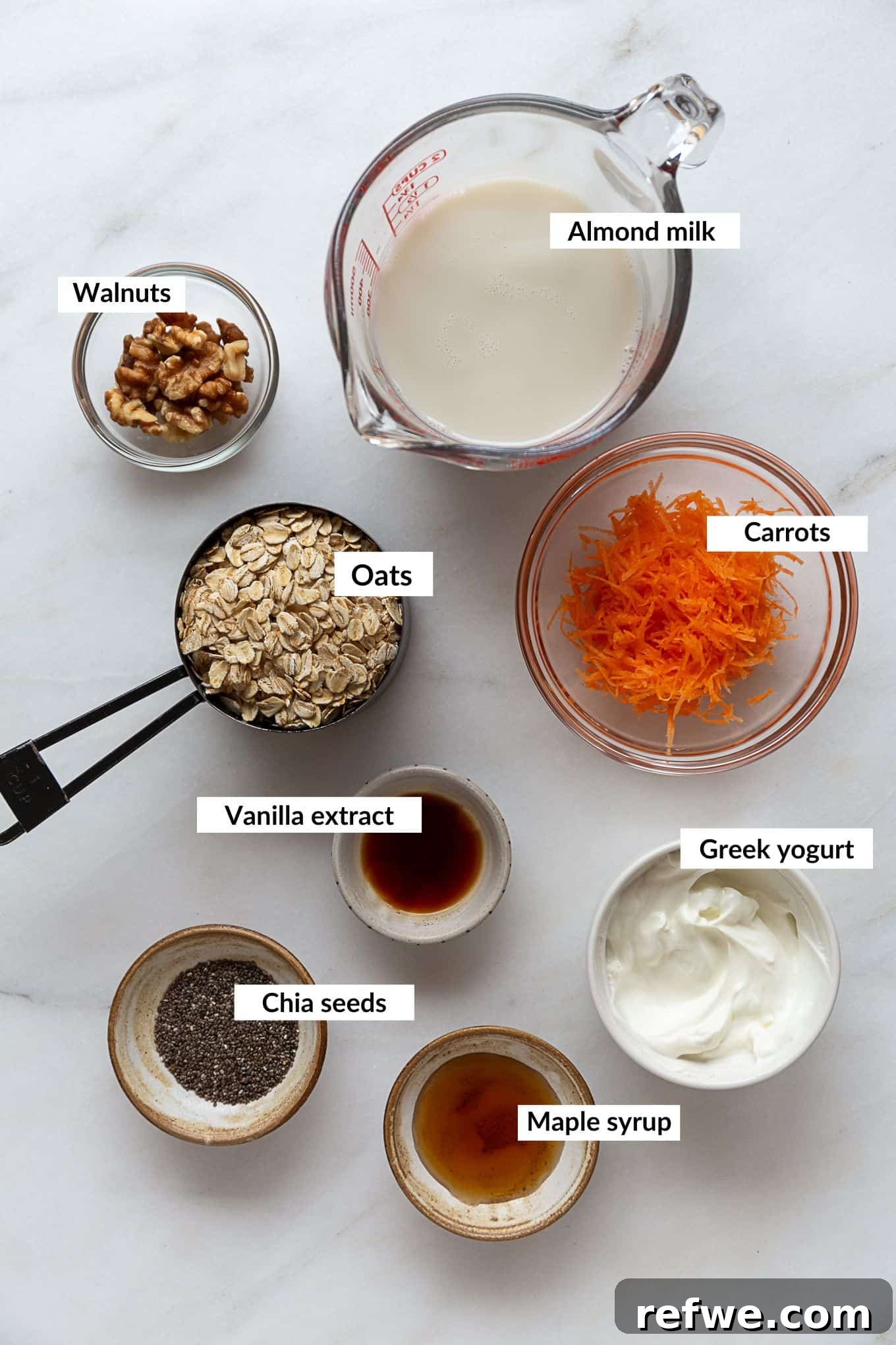 A flat lay of fresh ingredients for making carrot cake overnight oats, including oats, chia seeds, shredded carrots, cinnamon, nutmeg, yogurt, and almond milk.