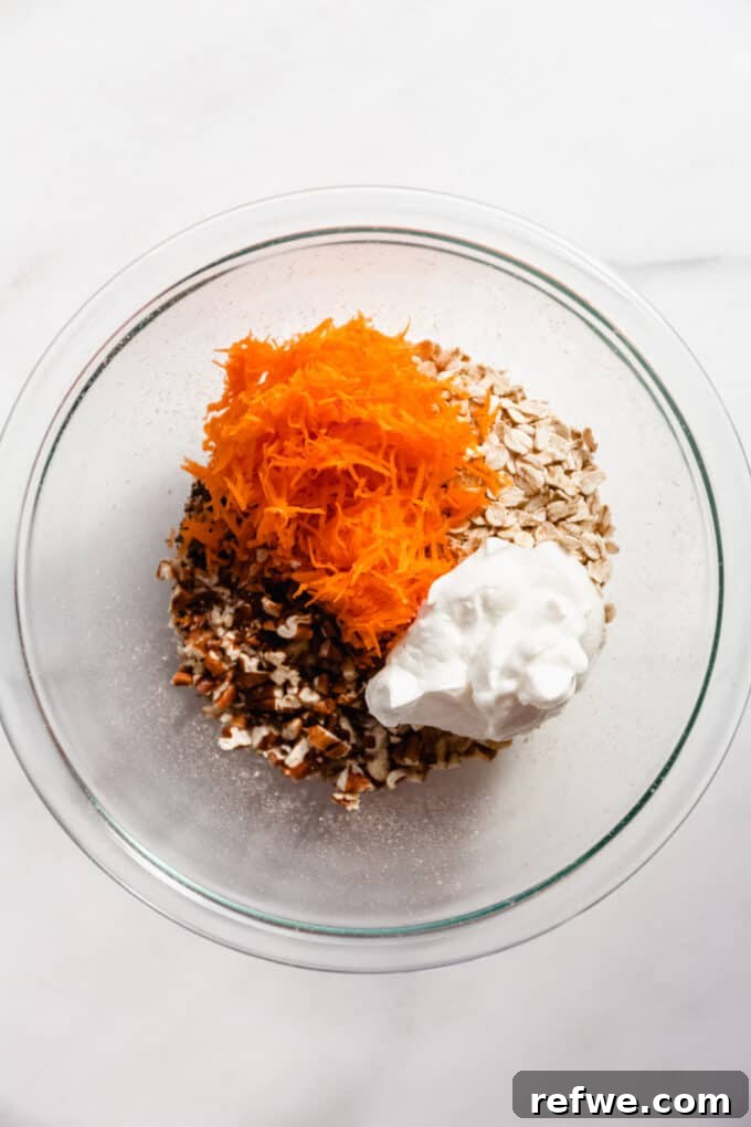 A clear mixing bowl filled with dry ingredients for carrot cake overnight oats, including oats, walnuts, yogurt, and shredded carrots, before mixing with liquid.