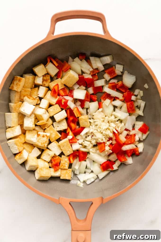 Effortless Kung Pao Tofu 5 A mix of crispy tofu, vibrant red peppers, chopped onions, and minced garlic simmering together in a pink pan.