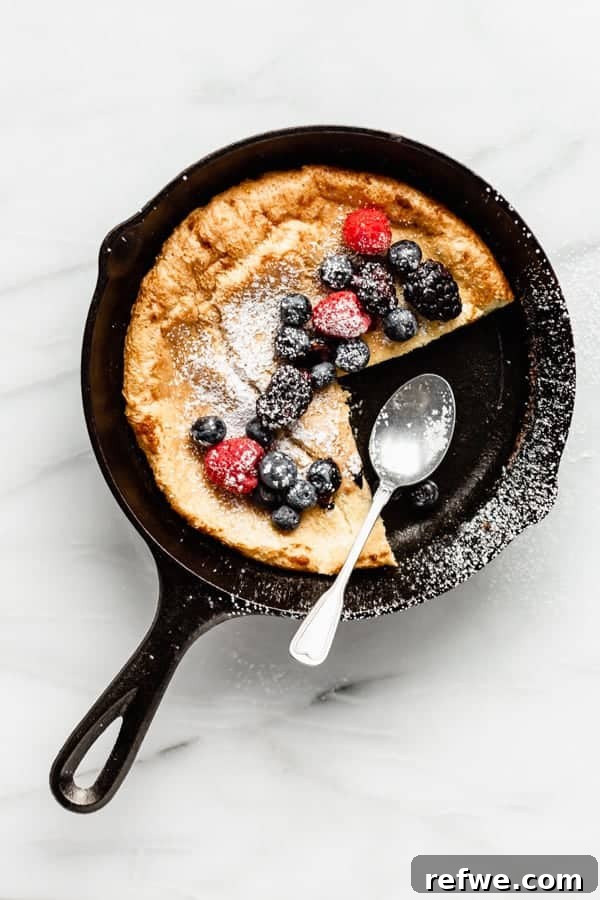 Puffed Skillet Pancake 2 A golden, fluffy Dutch Baby Pancake baked to perfection in a cast iron skillet, garnished with an array of vibrant fresh berries and a sprinkle of powdered sugar, with a serving spoon resting beside it.