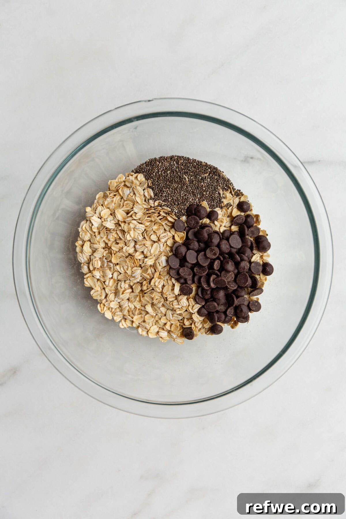Chocolate Chip Cookie Crunch Granola 4 A clear mixing bowl holds a blend of oats, chia seeds, and chocolate chips, ready for the next step.