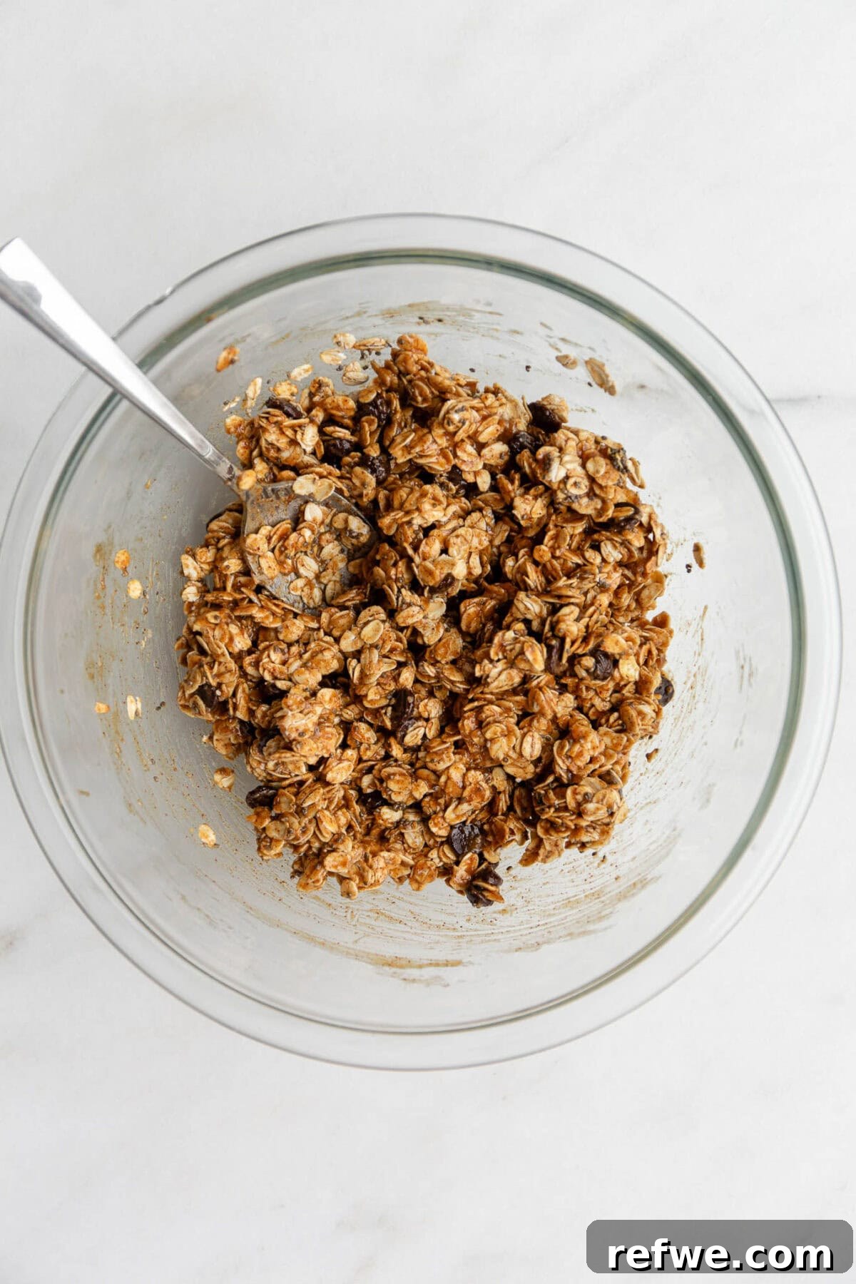 Chocolate Chip Cookie Crunch Granola 7 The chocolate chip cookie granola ingredients are thoroughly mixed in a clear bowl with a spatula, achieving an even coating.