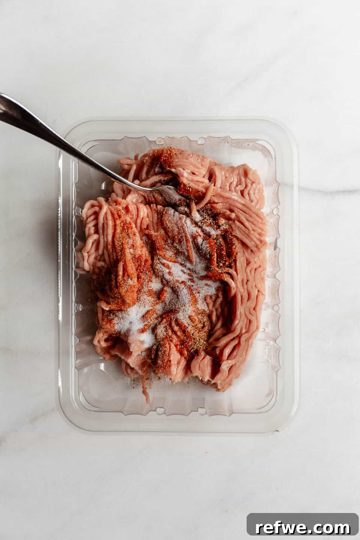 Raw ground chicken in a plastic container being seasoned with spices and mixed with a fork.