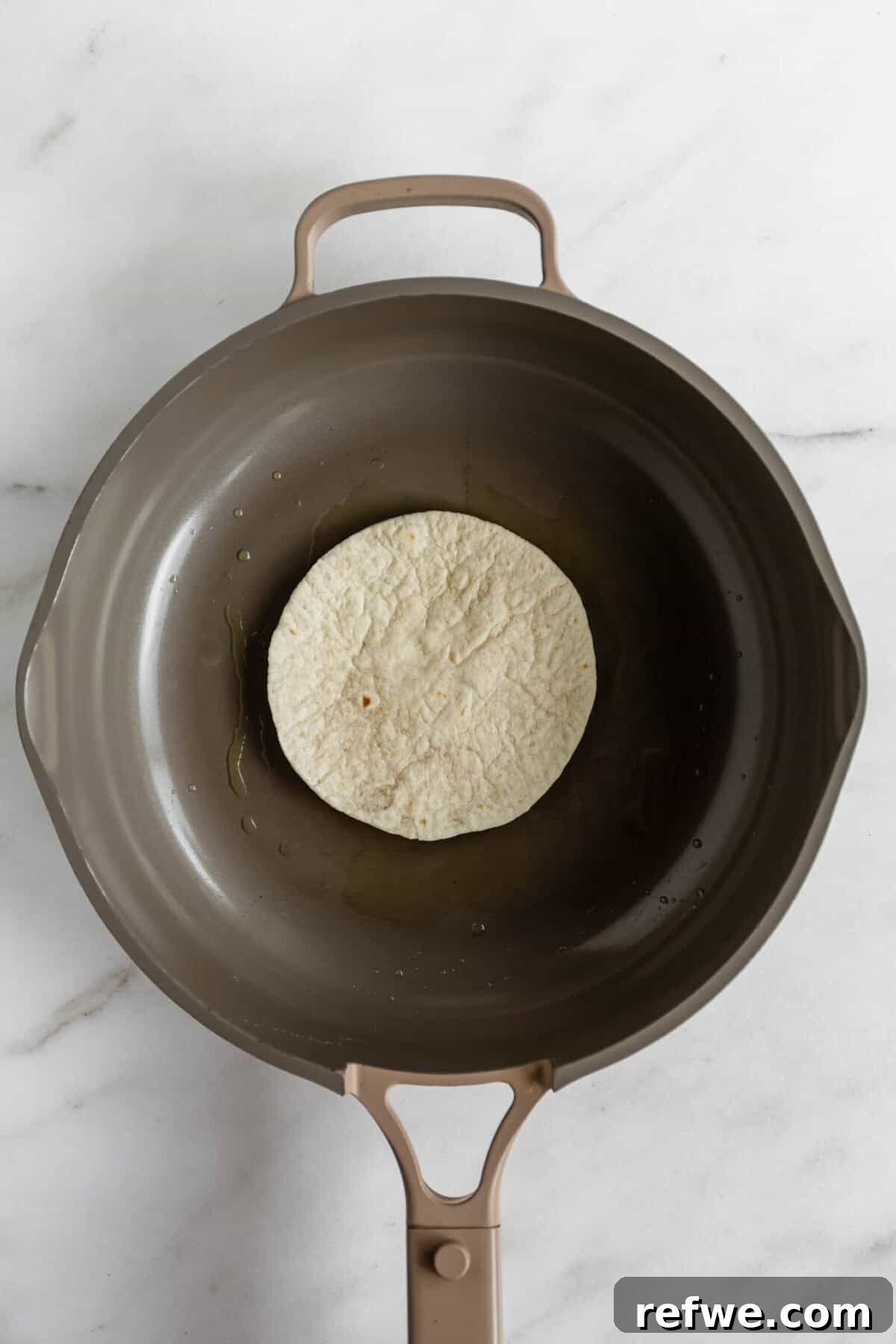 A flour tortilla in a dark skillet on a marble surface, ready to be toasted for chicken caesar smashed tacos.