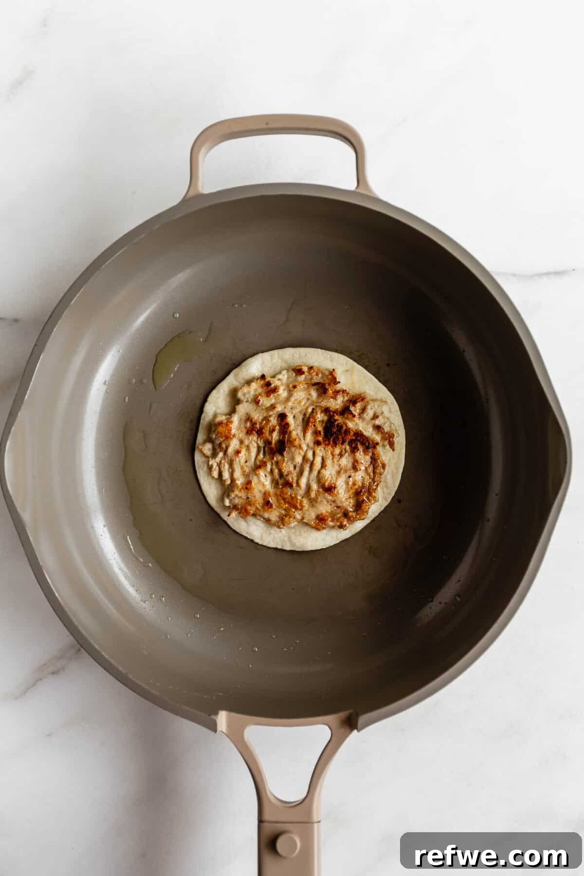 A toasted smashed tortilla in a dark skillet viewed from above, showing a golden brown crispy base for chicken caesar smashed tacos.
