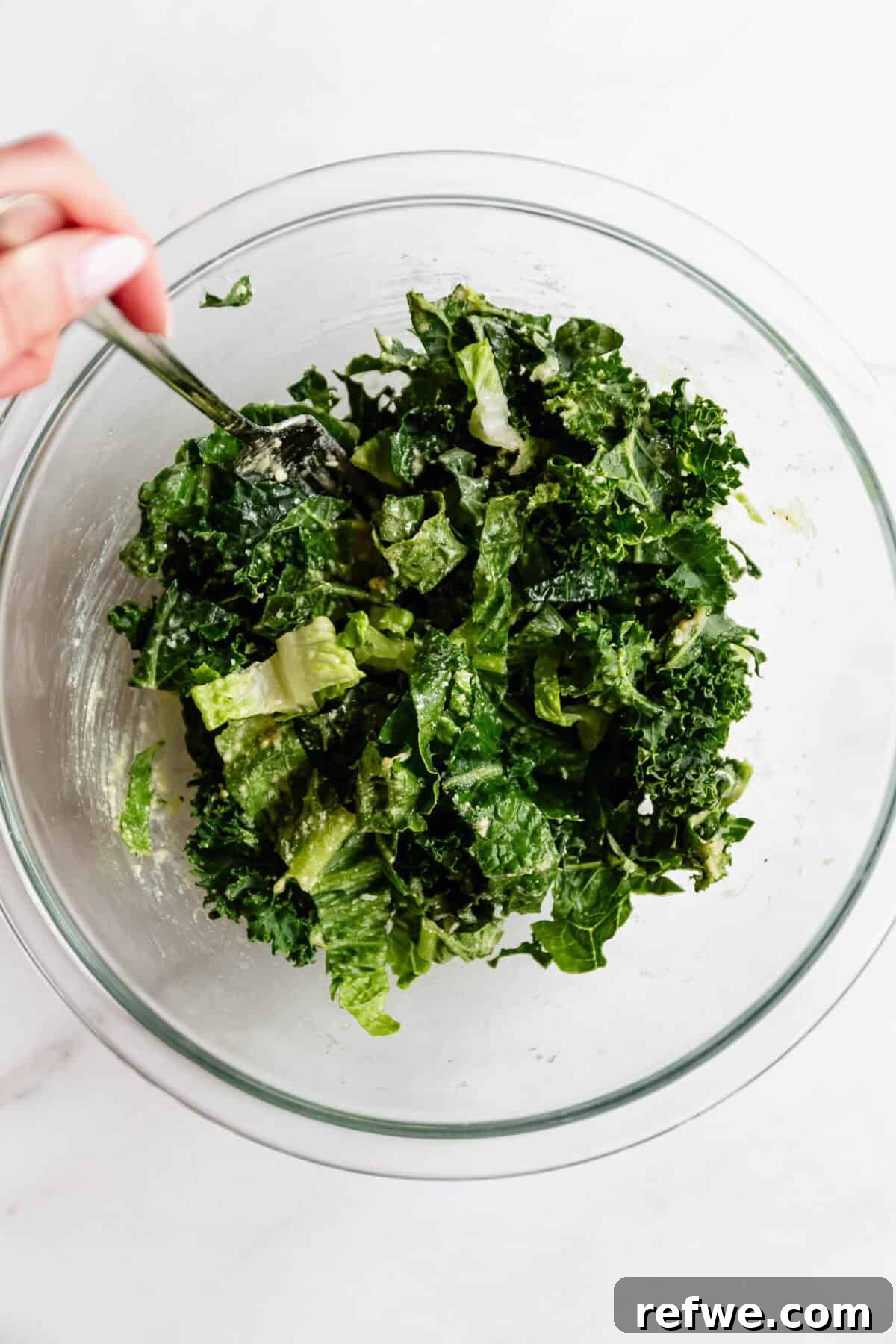 Hand tossing kale caesar salad with tongs in a glass bowl to prepare the topping for chicken caesar smashed tacos.