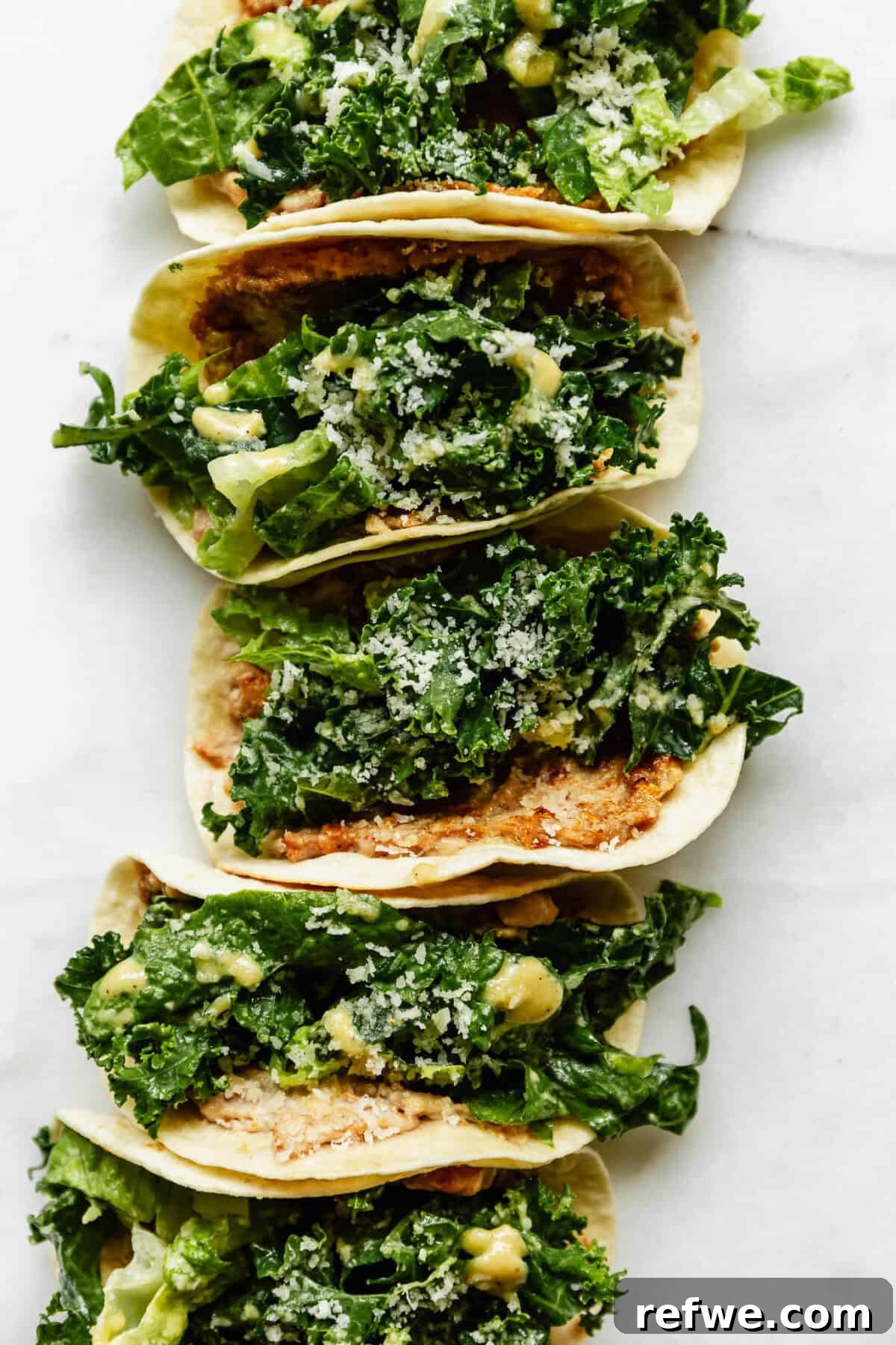 Three chicken caesar smashed tacos arranged in a row on white marble, topped with kale caesar salad and parmesan cheese.