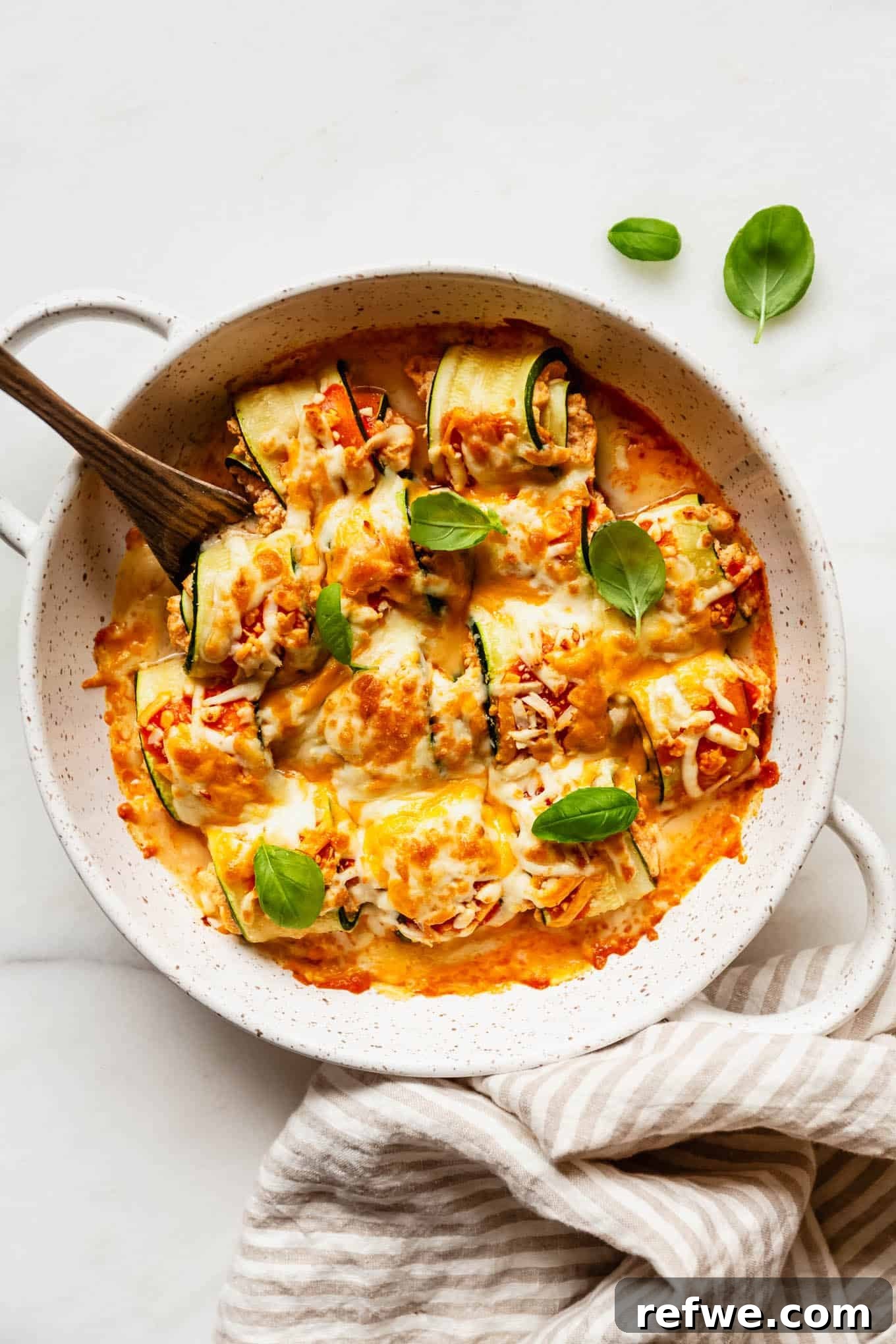 Buffalo Chicken Zucchini Roll-Ups Recipe 2 buffalo chicken zucchini roll ups in a baking dish with a beige stripe napkin on the side.