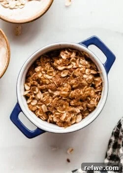 Unbaked apple crisp in a small blue ramekin surrounded by bowls of oats on a marble counter.