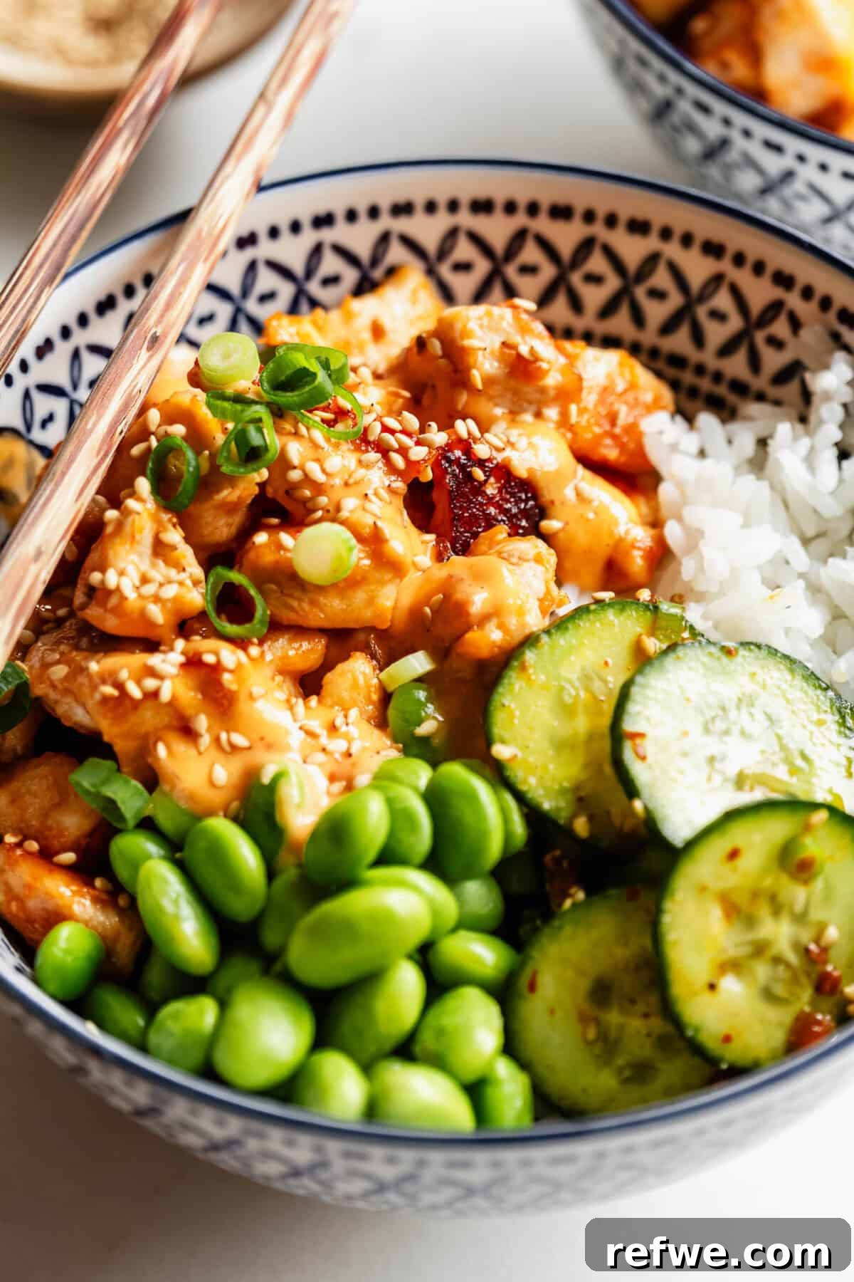 Bang Bang Chicken Bowl Recipe with Spicy Cucumber Salad 11 Close-up of a bang bang chicken bowl with rice, cucumber slices, edamame, and chopsticks resting on top.
