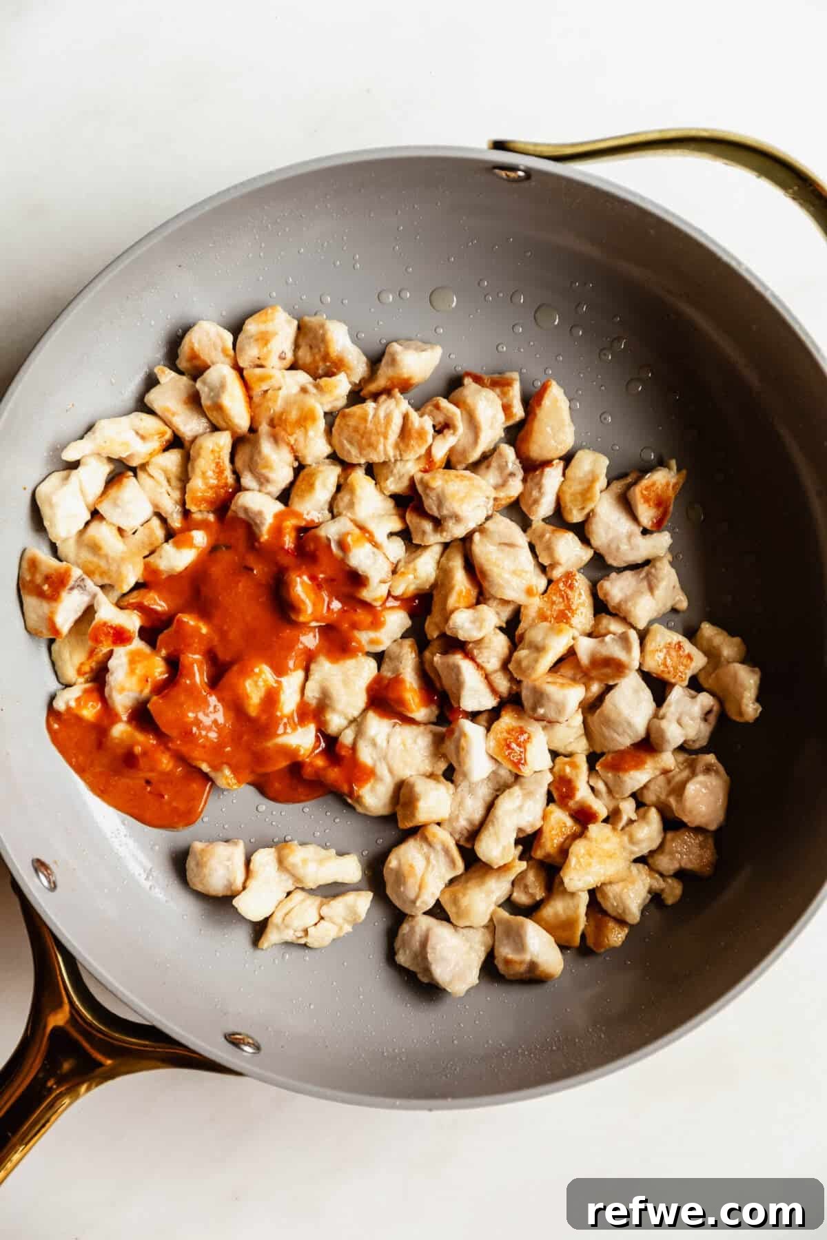Bang Bang Chicken Bowl Recipe with Spicy Cucumber Salad 6 Diced raw chicken pieces searing in a skillet with bang bang sauce being poured over half of the pan.